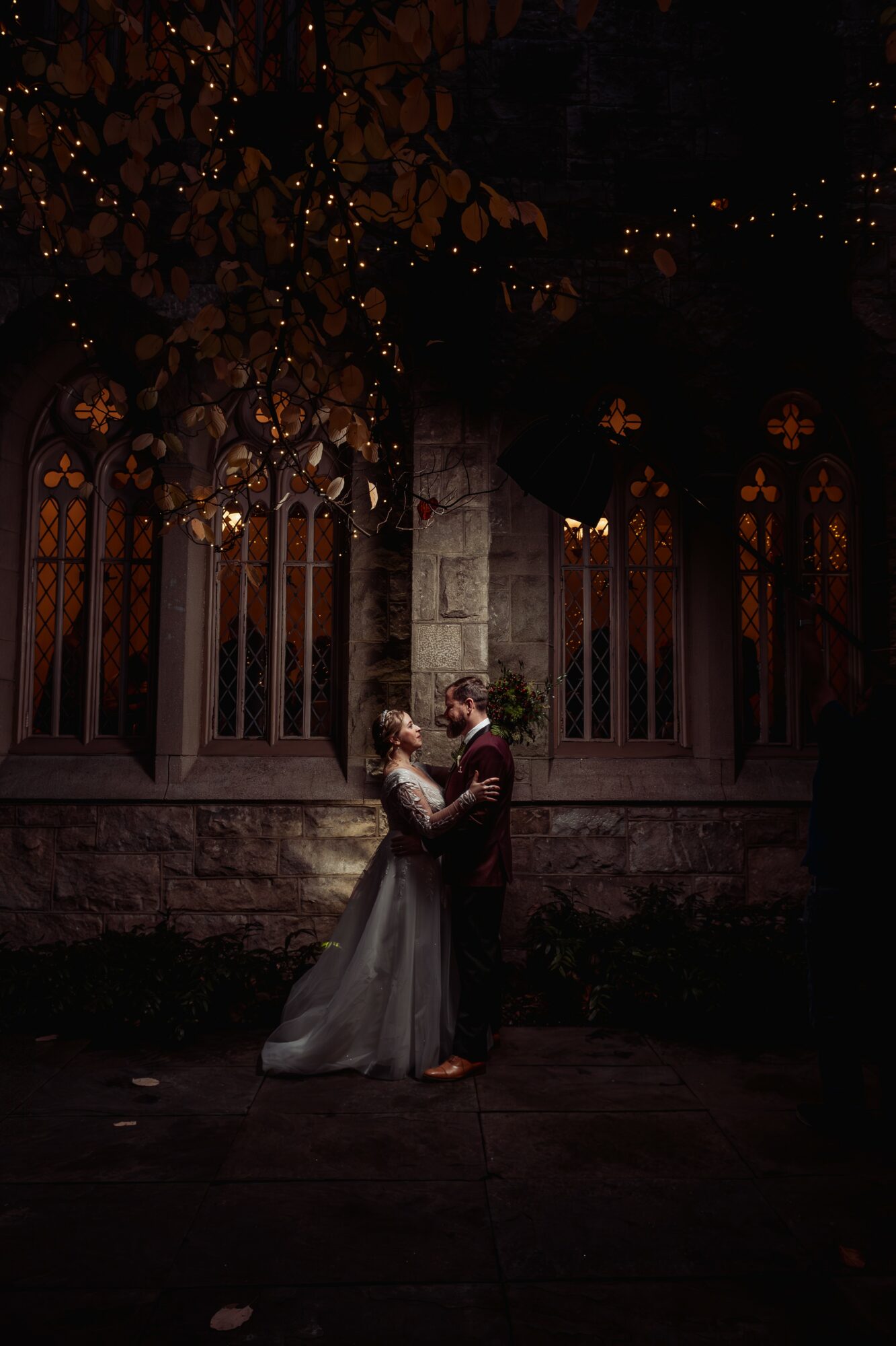 Couple dancing outdoors at night in front of a stone wall with tall arched windows and trees with lights.