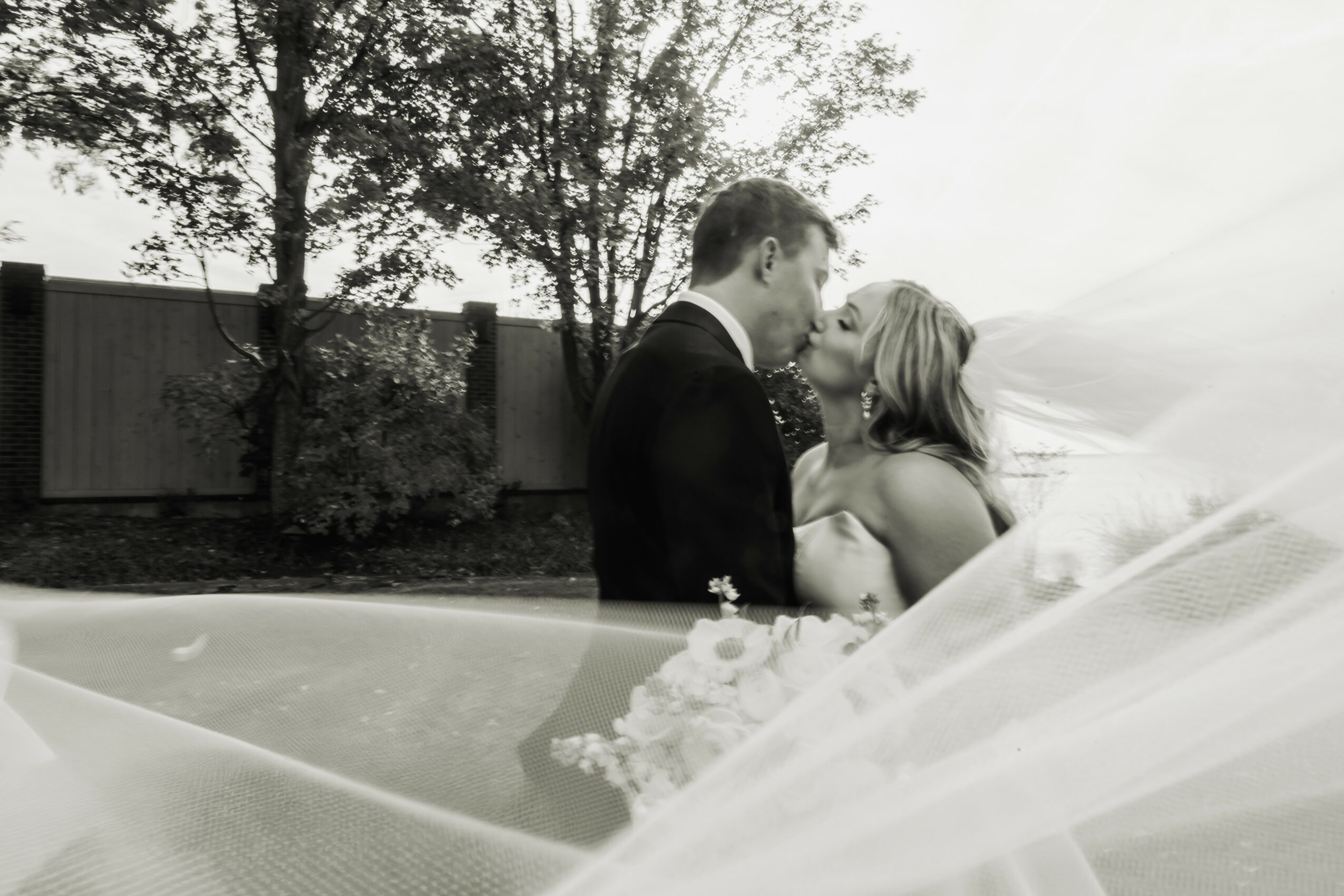 A couple kissing outdoors with trees and a fence in the background, black and white photo, wedding attire, veil visible.