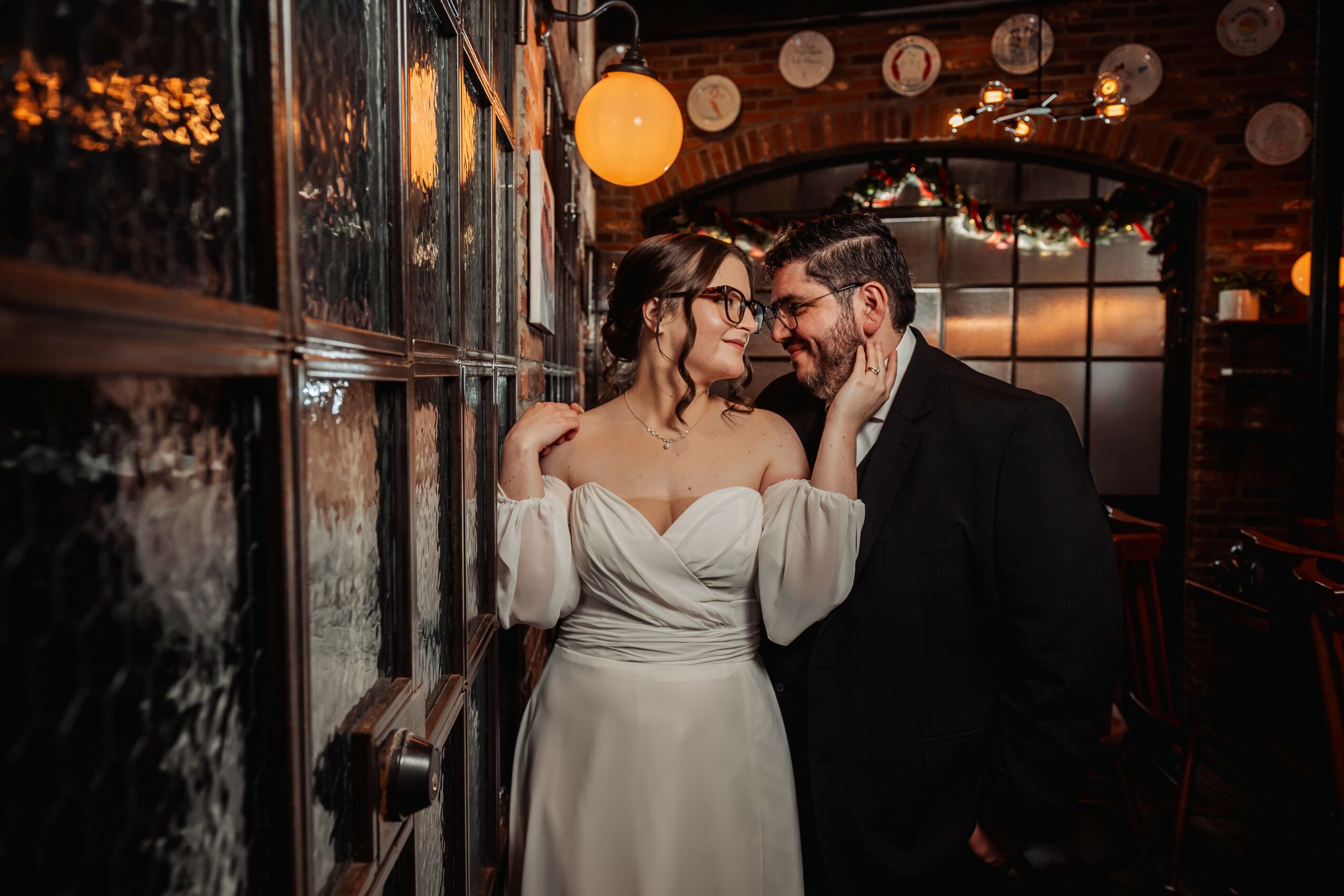 A couple dressed in formal attire close together indoors, smiling and looking at each other, with warm lighting and wooden decor.