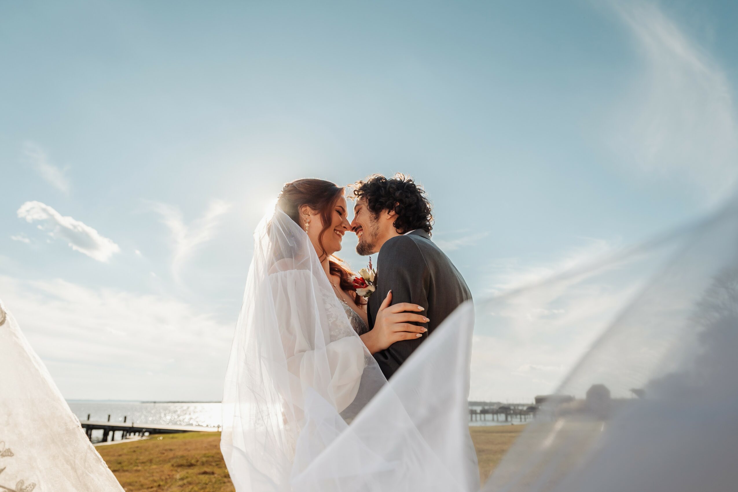 Bride and groom face each other outdoors, with sky and clouds in the background, close together, smiling.