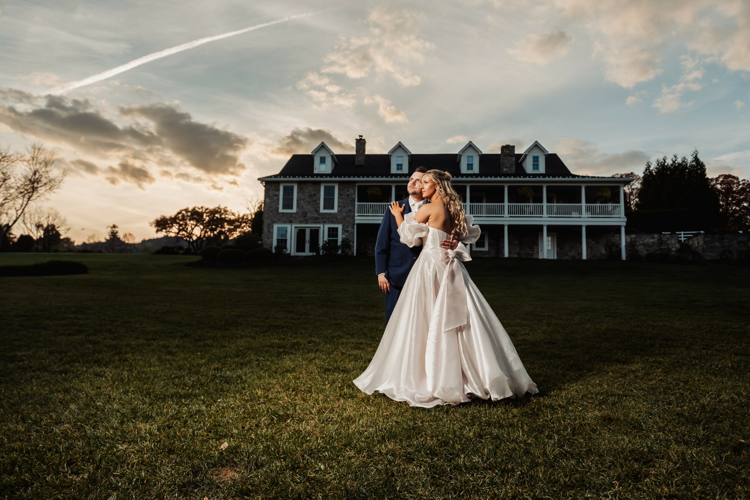 Bride and groom standing on grass in front of large house during sunset, embracing each other.