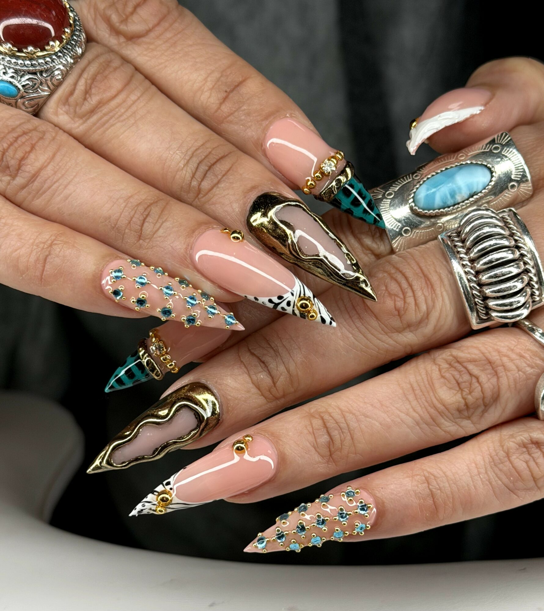 Close-up of hands with elaborate, decorated nails and multiple rings, featuring gold, turquoise, and rhinestone accents.