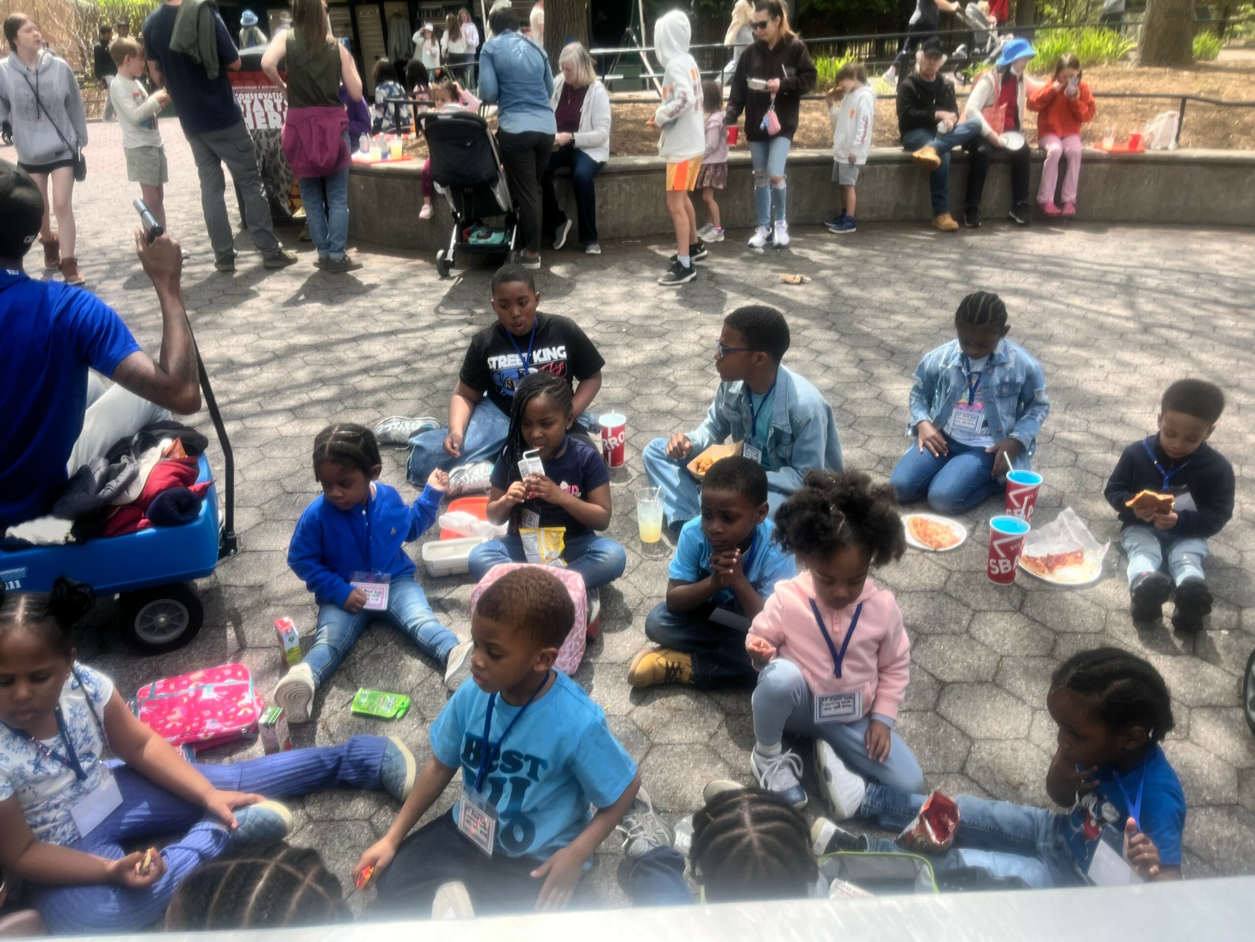 Children sitting on the ground with adults and other children in the background outdoors.