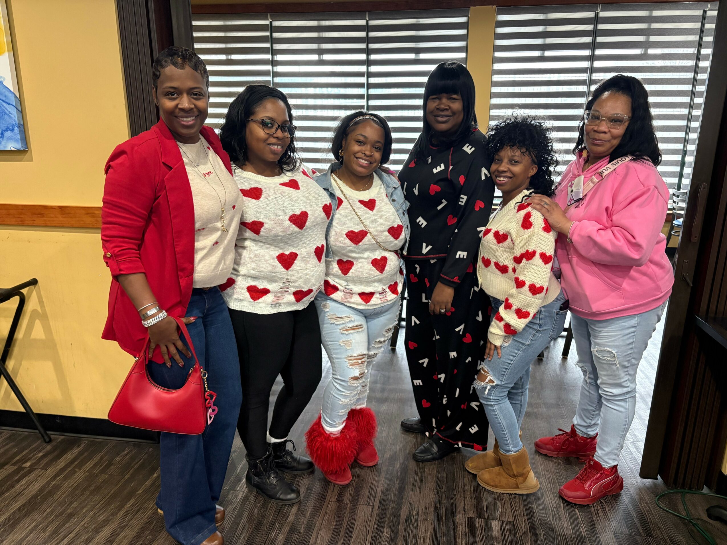 Six women standing indoors, smiling, wearing casual clothes with heart patterns and colorful footwear.