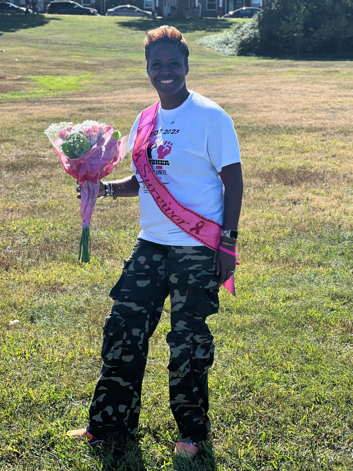 Person standing outdoors on grass holding a bouquet of flowers, smiling, wearing a white t-shirt and camouflage pants.