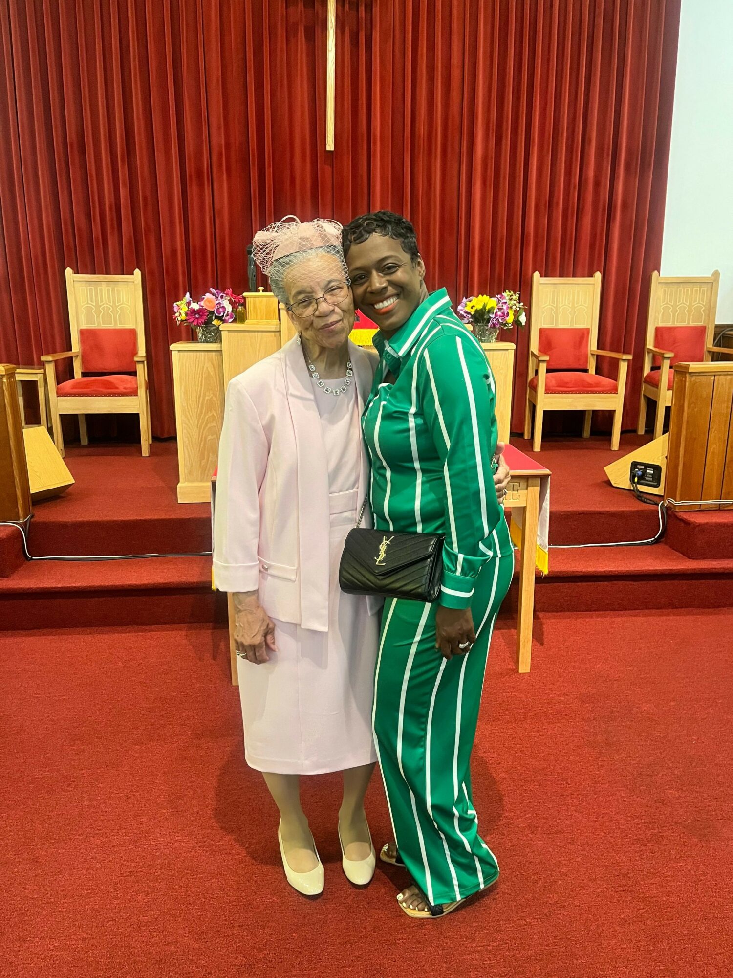 Two women standing together in a room with red curtains and chairs, smiling, one in a light-colored outfit, the other in green striped pajamas.