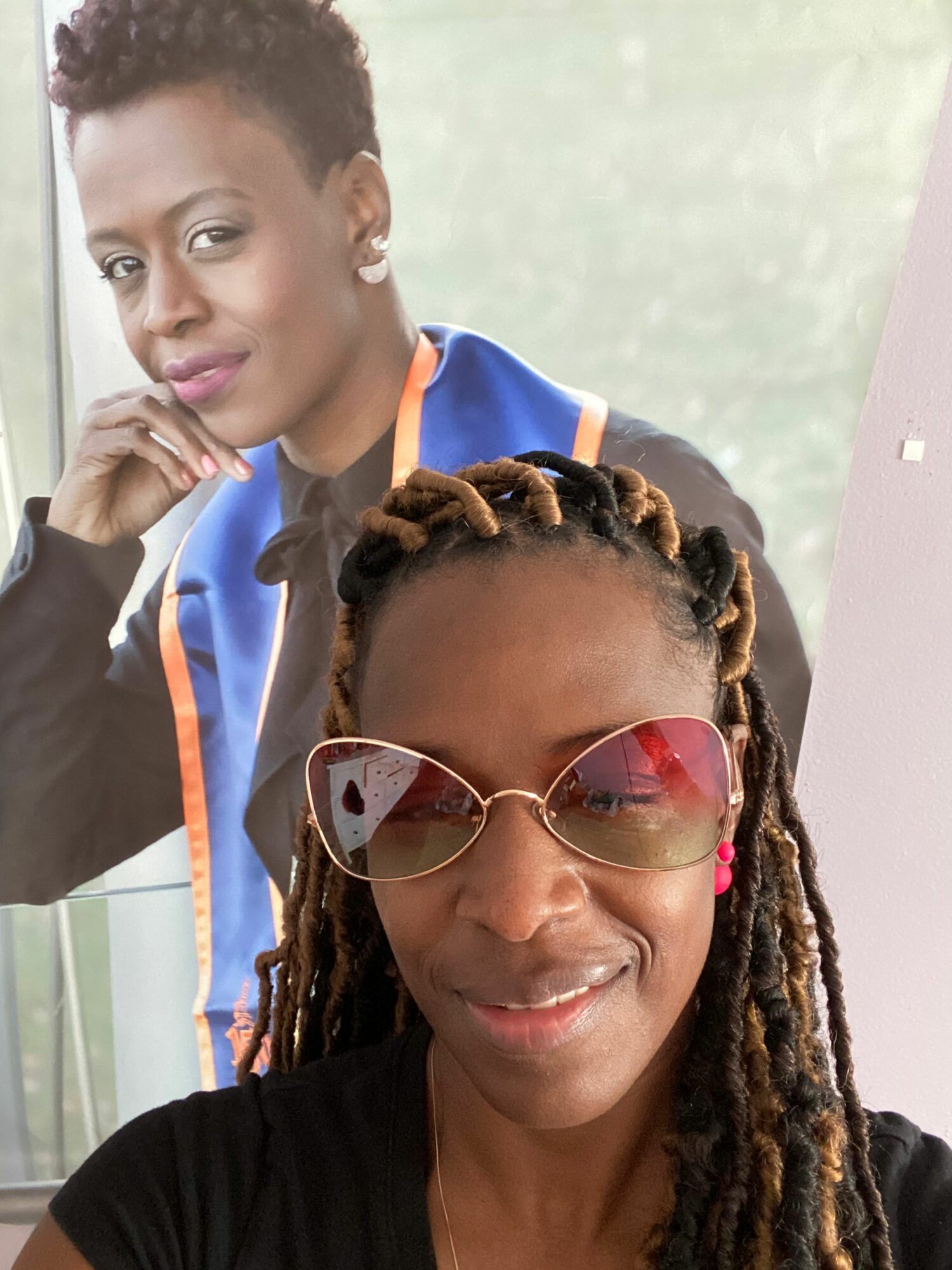 Two women pose for a selfie indoors, one wearing sunglasses and the other with short curly hair, both smiling.