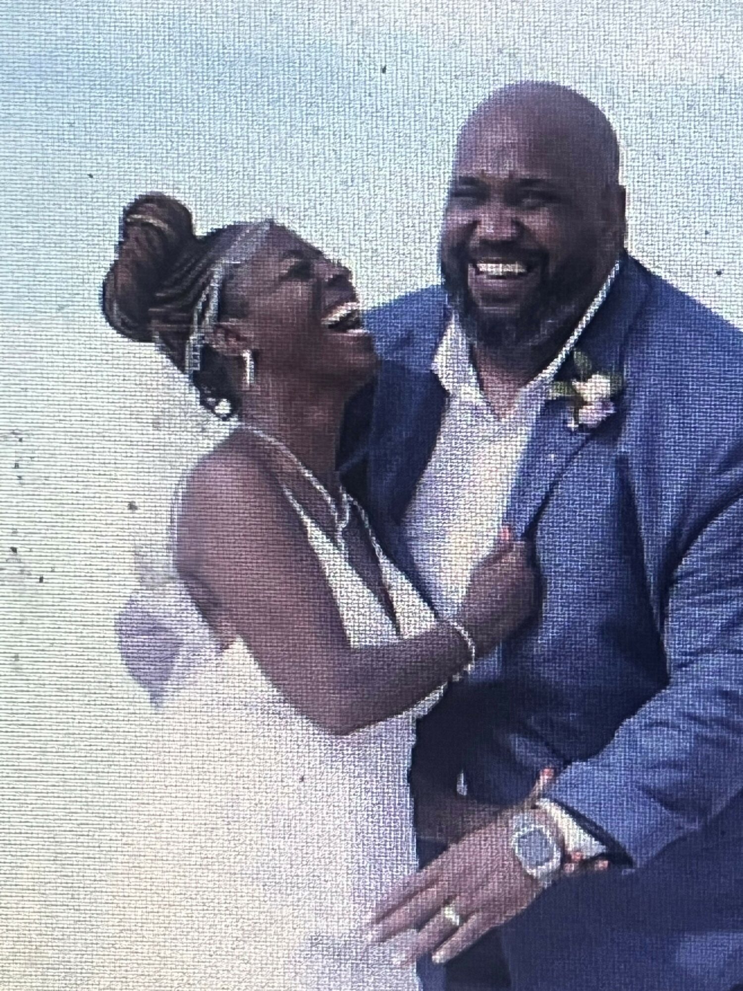 A joyful couple dressed in formal attire, smiling and embracing outdoors against a light background.