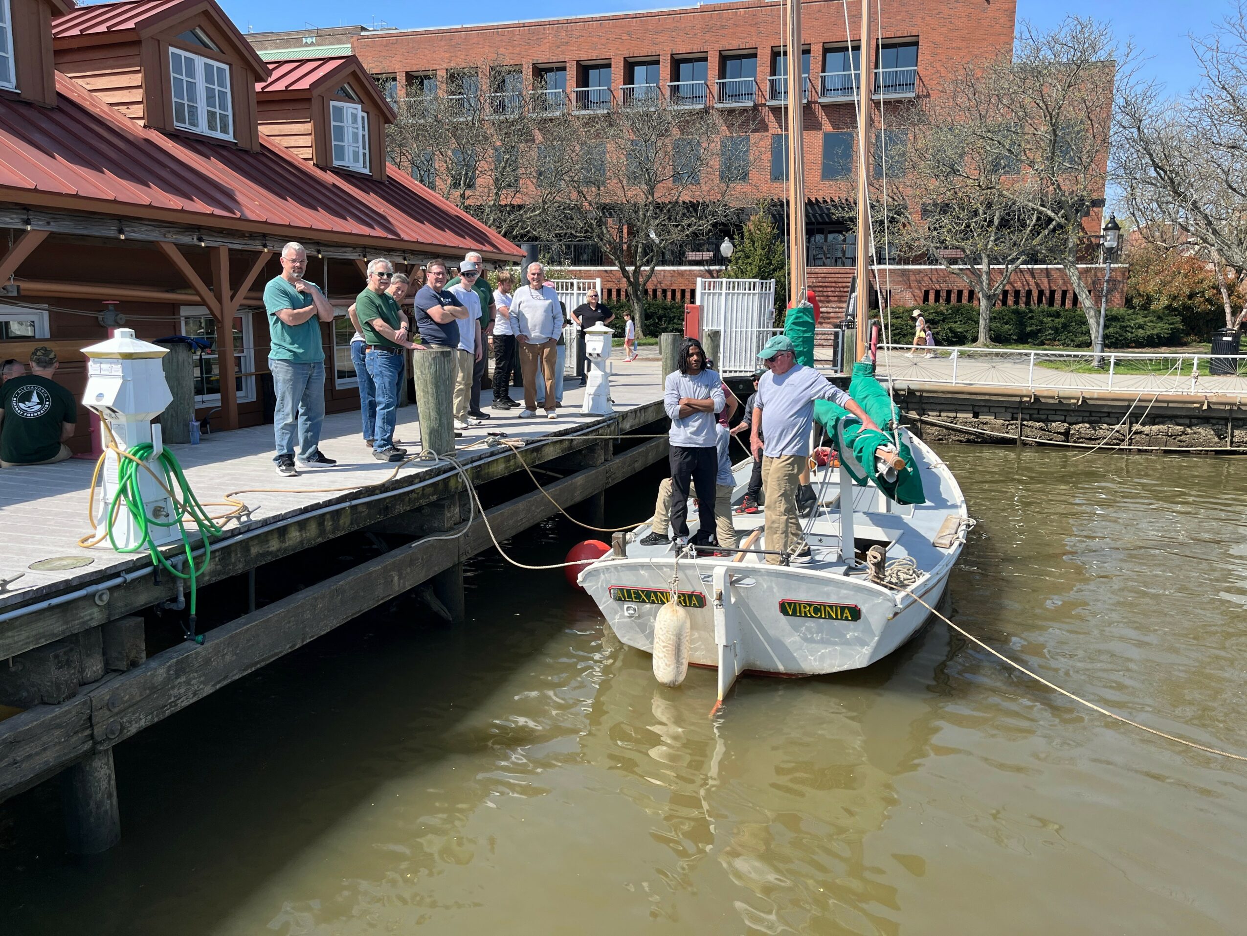 Group of people on dock near boat, some holding ropes, with building and trees in background.