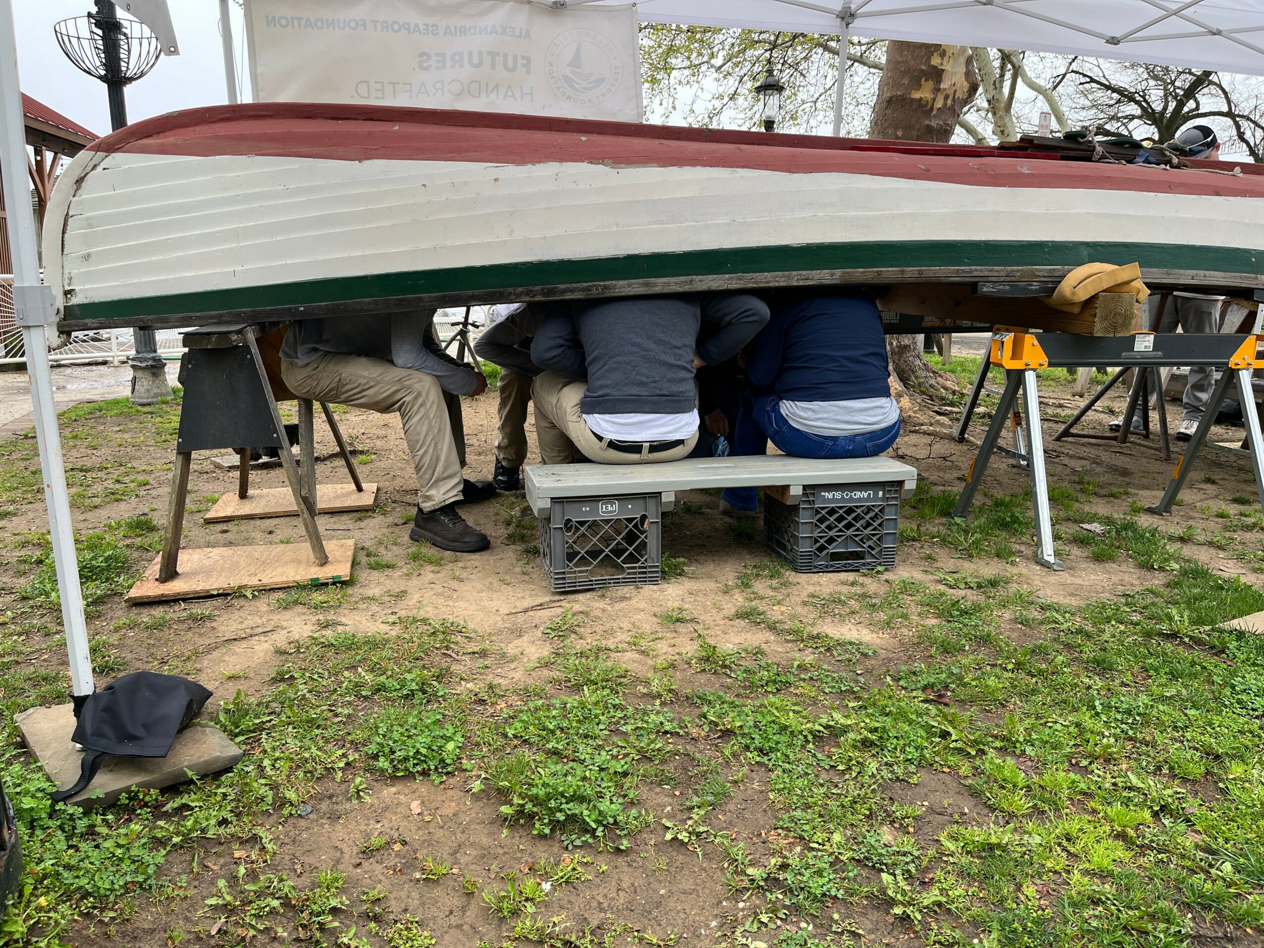 Three people sitting under a boat on a grassy area, with tools and equipment nearby.