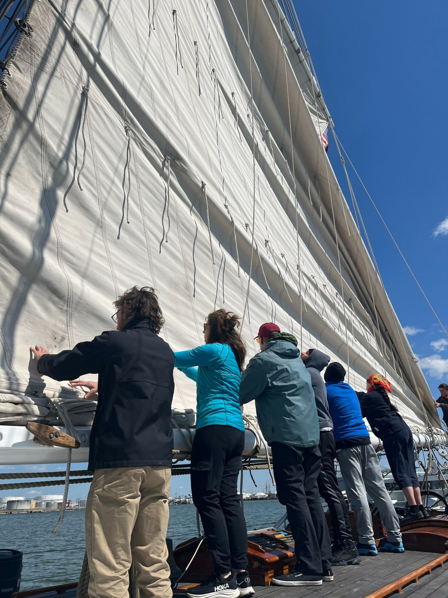 Group of people standing on a dock, looking at a large sailboat with a white sail, under a blue sky.