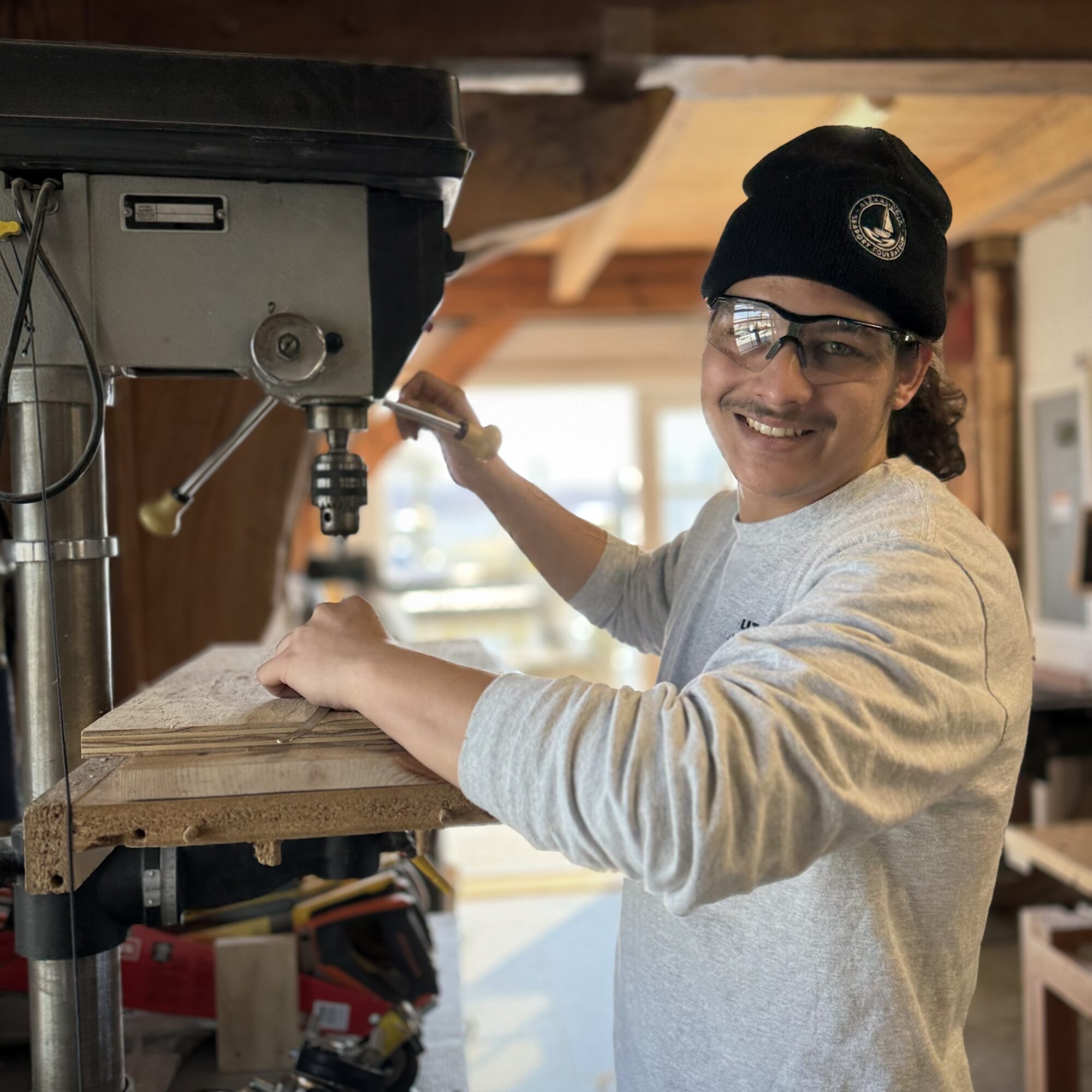 Person smiling, wearing glasses and a black beanie, working with a drill in a woodworking shop.