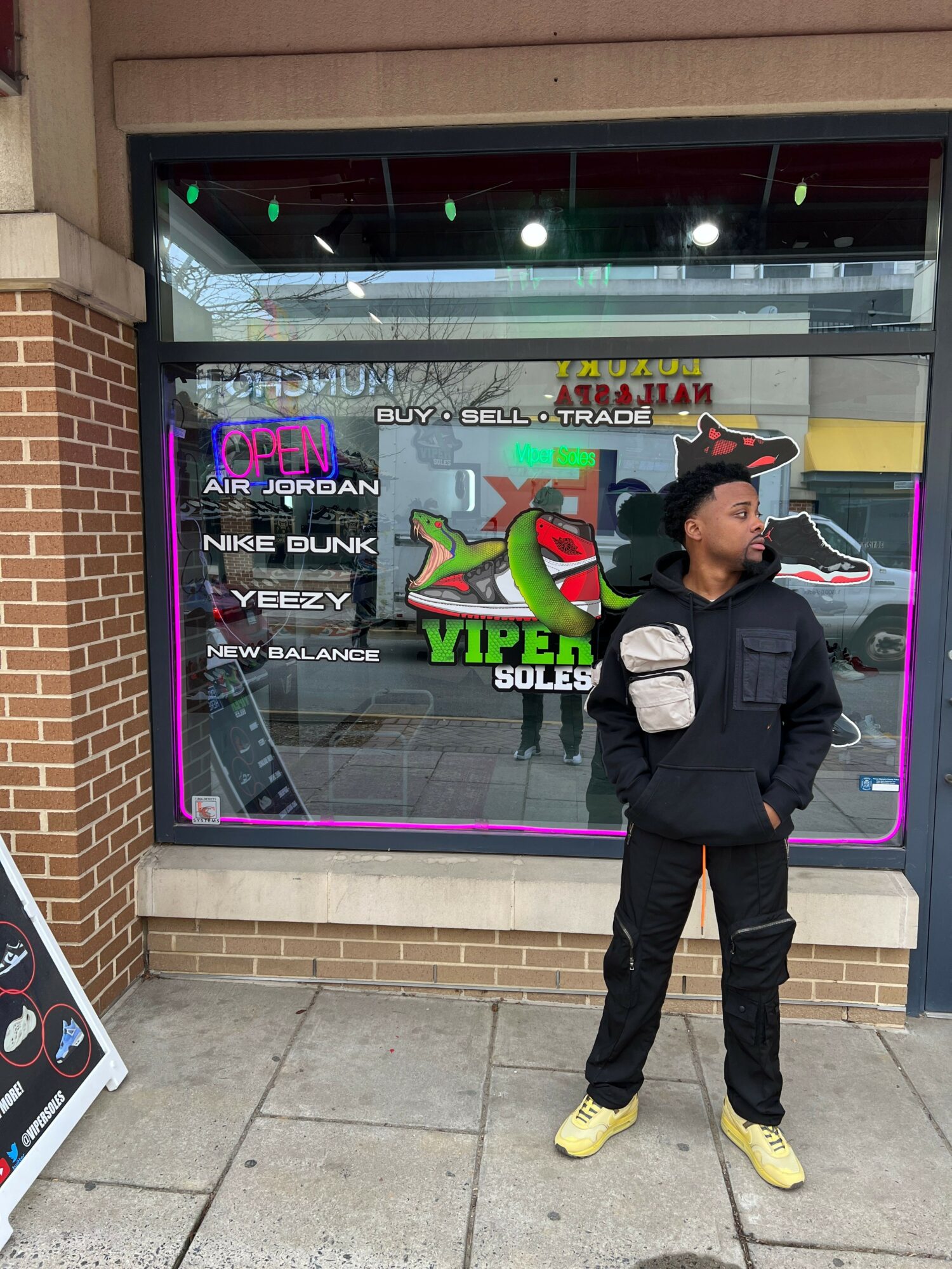 Young man in black jacket and yellow shoes standing outside store window with colorful signs and reflections.
