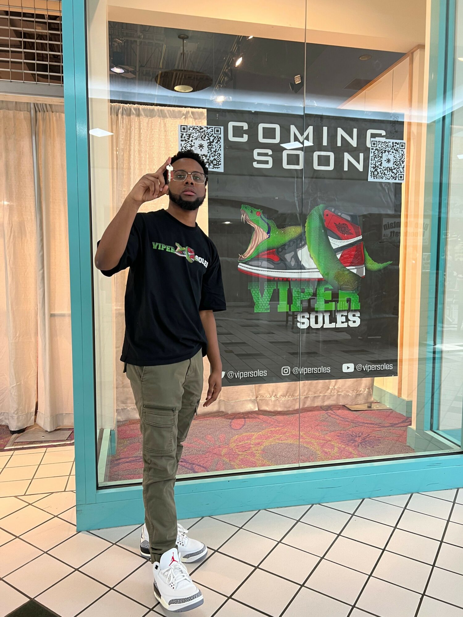 Young man standing inside store window, taking selfie, wearing black t-shirt, beige pants, white sneakers, with store sign behind him.