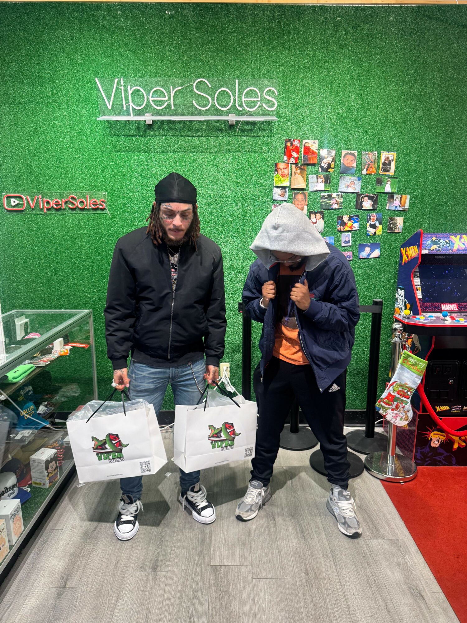 Two people standing in front of a green wall with store signage, holding shopping bags, with toys and arcade game nearby.