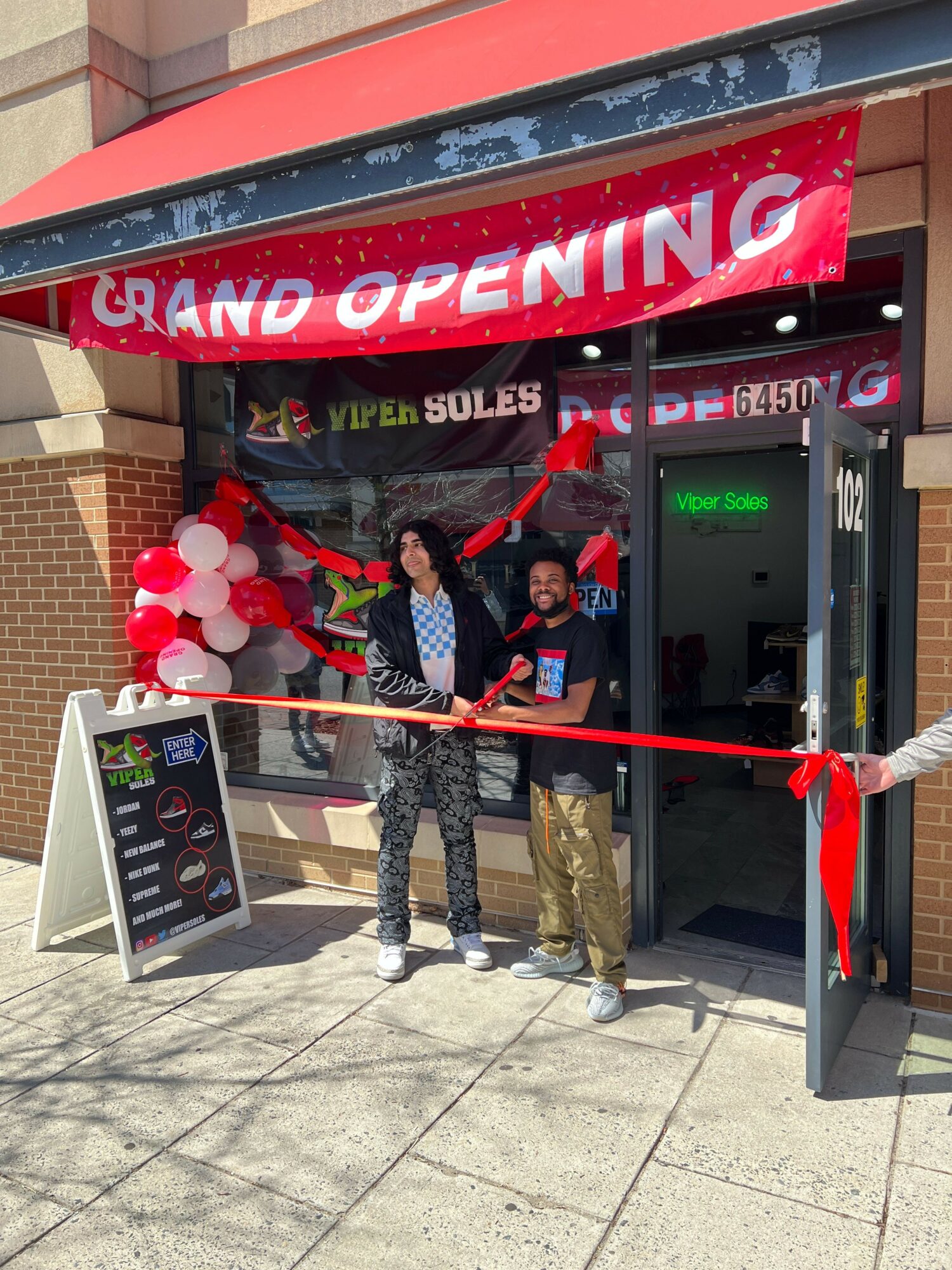 Two people stand outside a store with a red ribbon, balloons, and a grand opening banner, holding scissors for a ribbon-cutting ceremony.