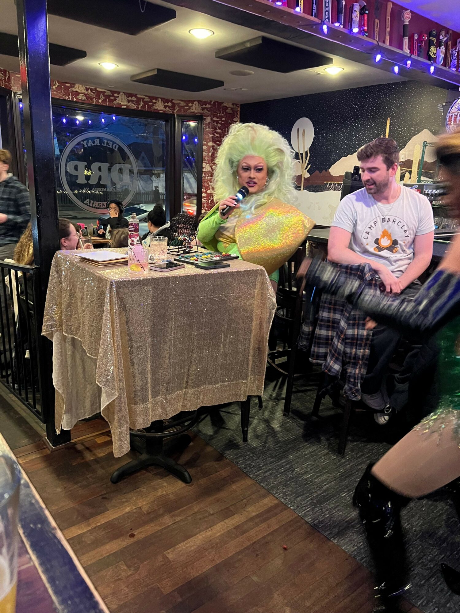 Person dressed in colorful costume with large white wig sitting at a table in a decorated room with string lights.
