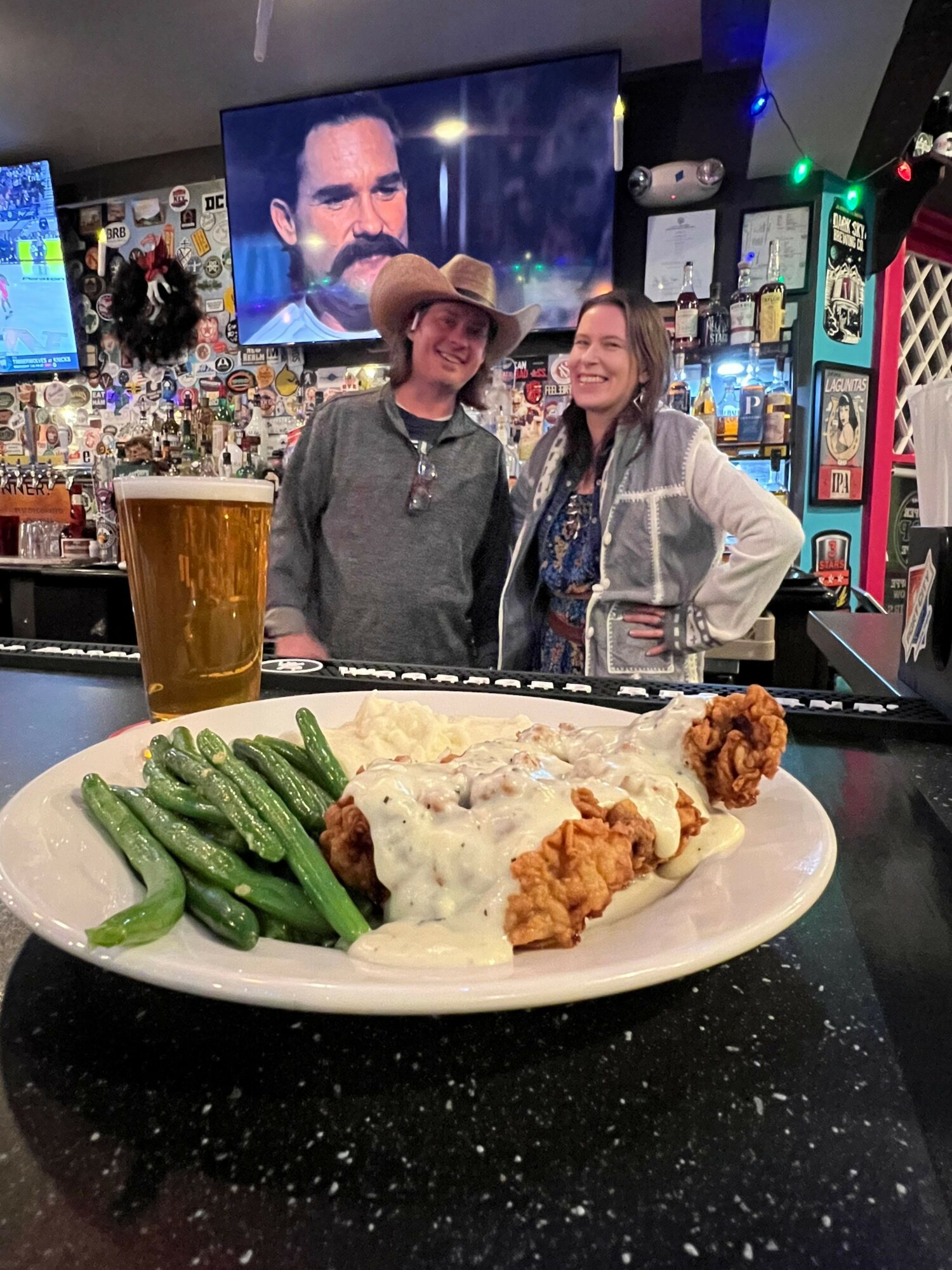 Two women smiling at a bar, with a plate of food including fried chicken, mashed potatoes, and green beans in foreground.