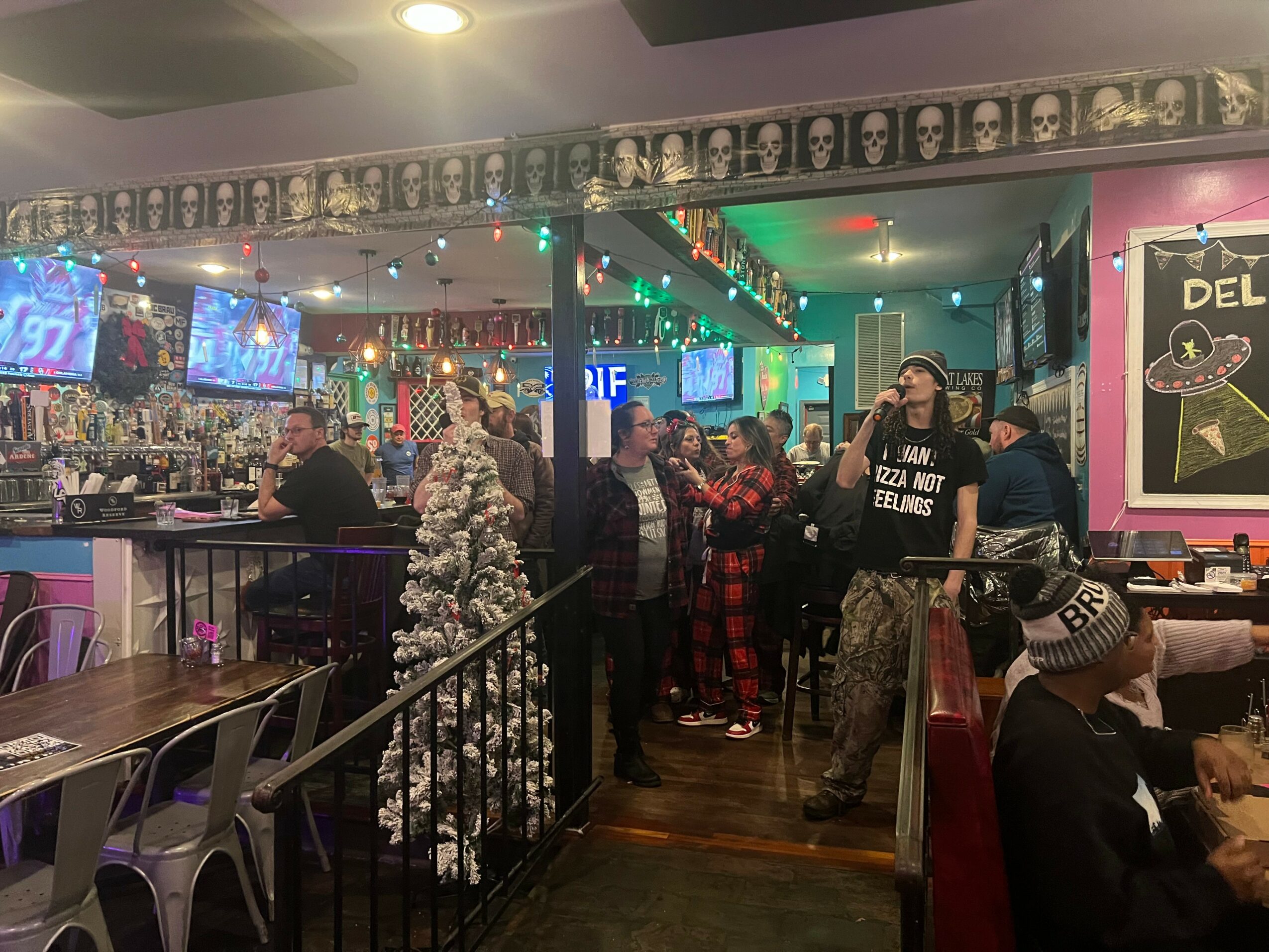 People gather in a decorated restaurant with Christmas trees, colorful lights, and wall art, some sitting and others standing.