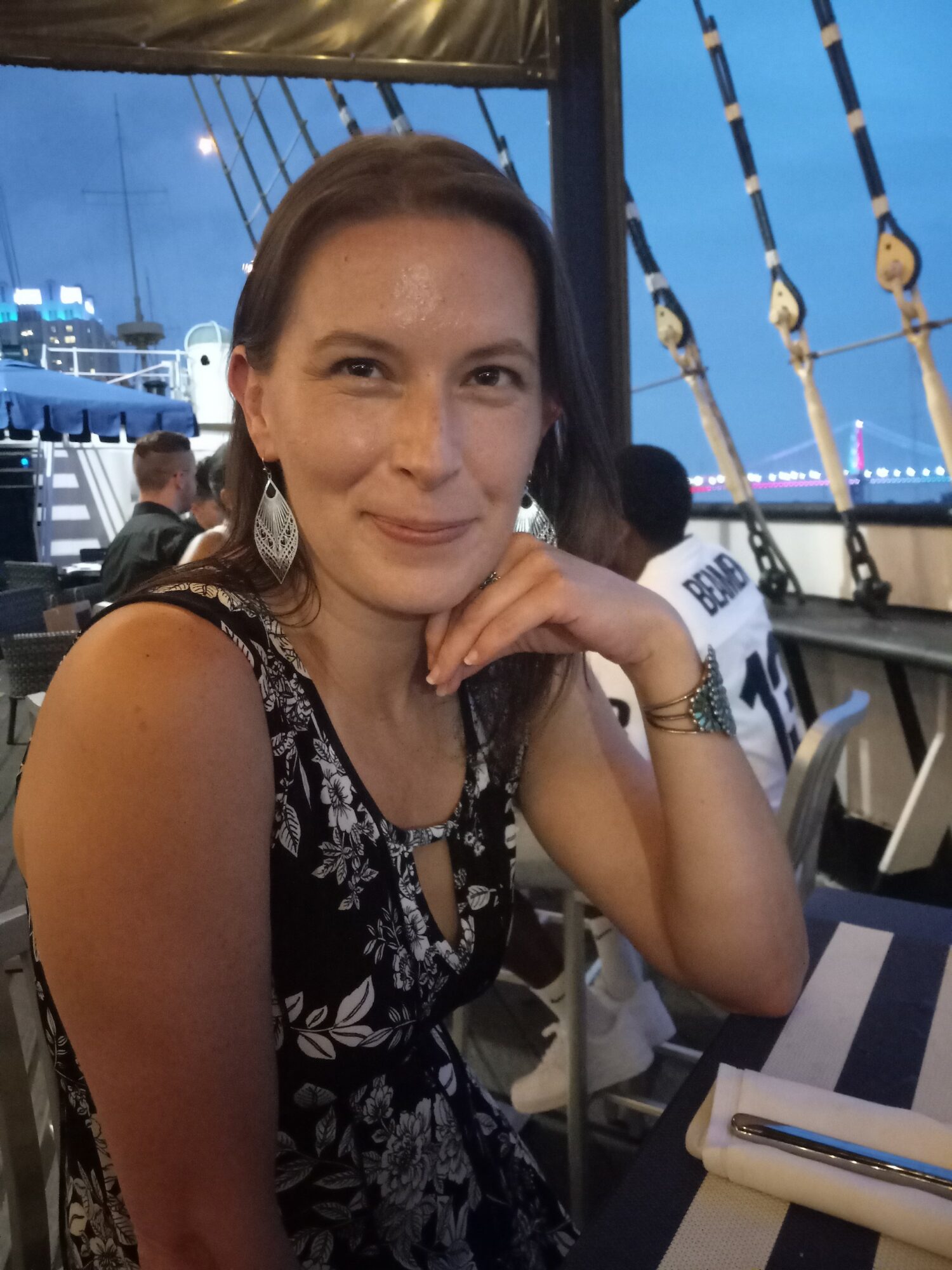 Woman with dark hair wearing a floral dress, sitting at a table outdoors near a boat, smiling.