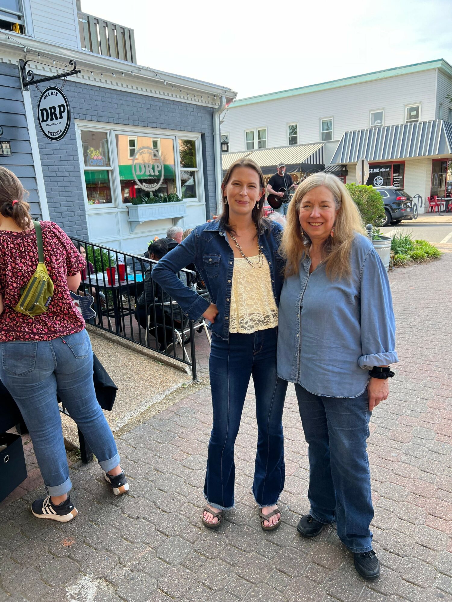Two women standing outside a building, smiling, with other people nearby and a parking lot in the background.
