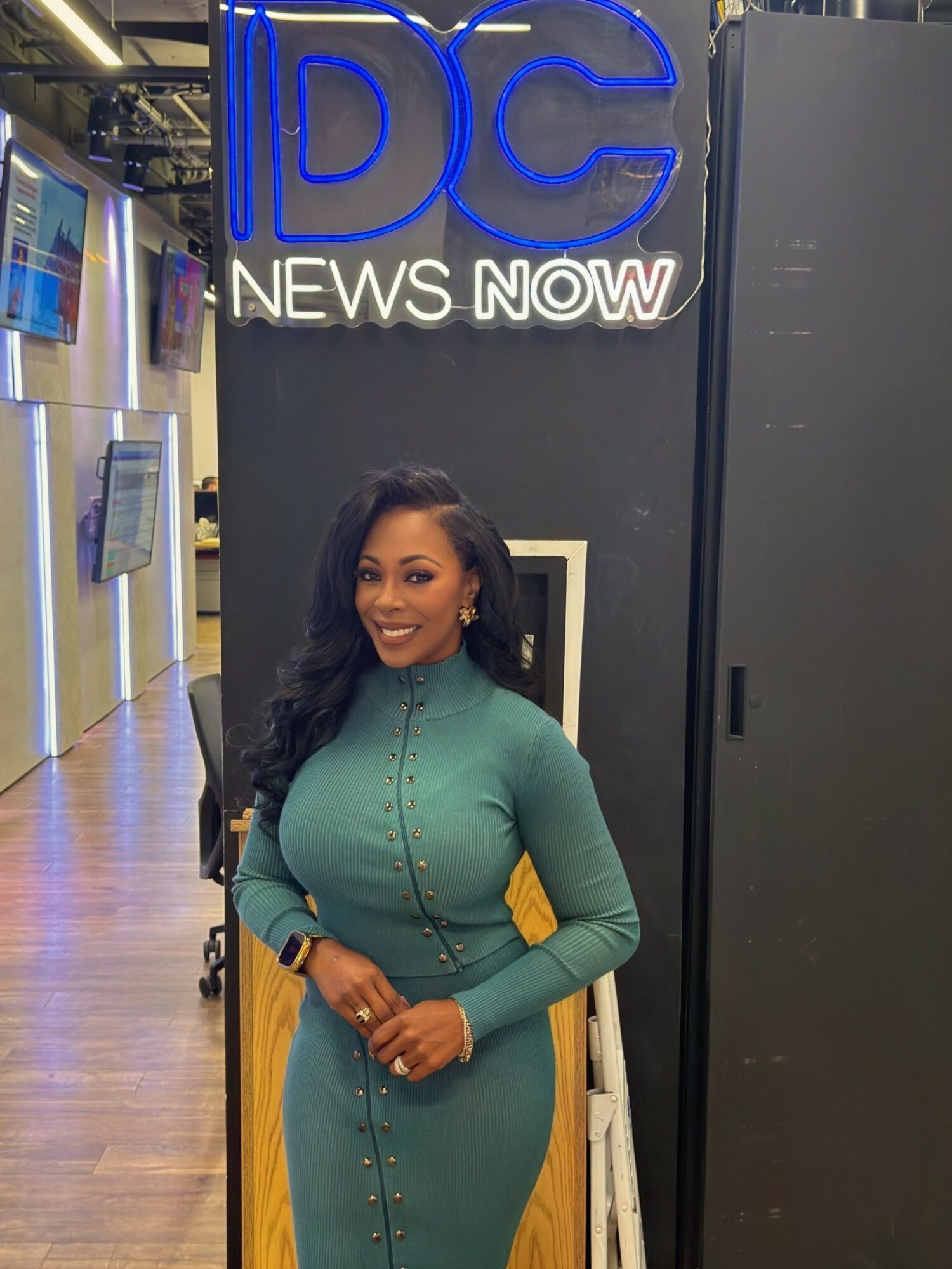 Woman with long dark hair in teal dress standing in front of a news station sign.