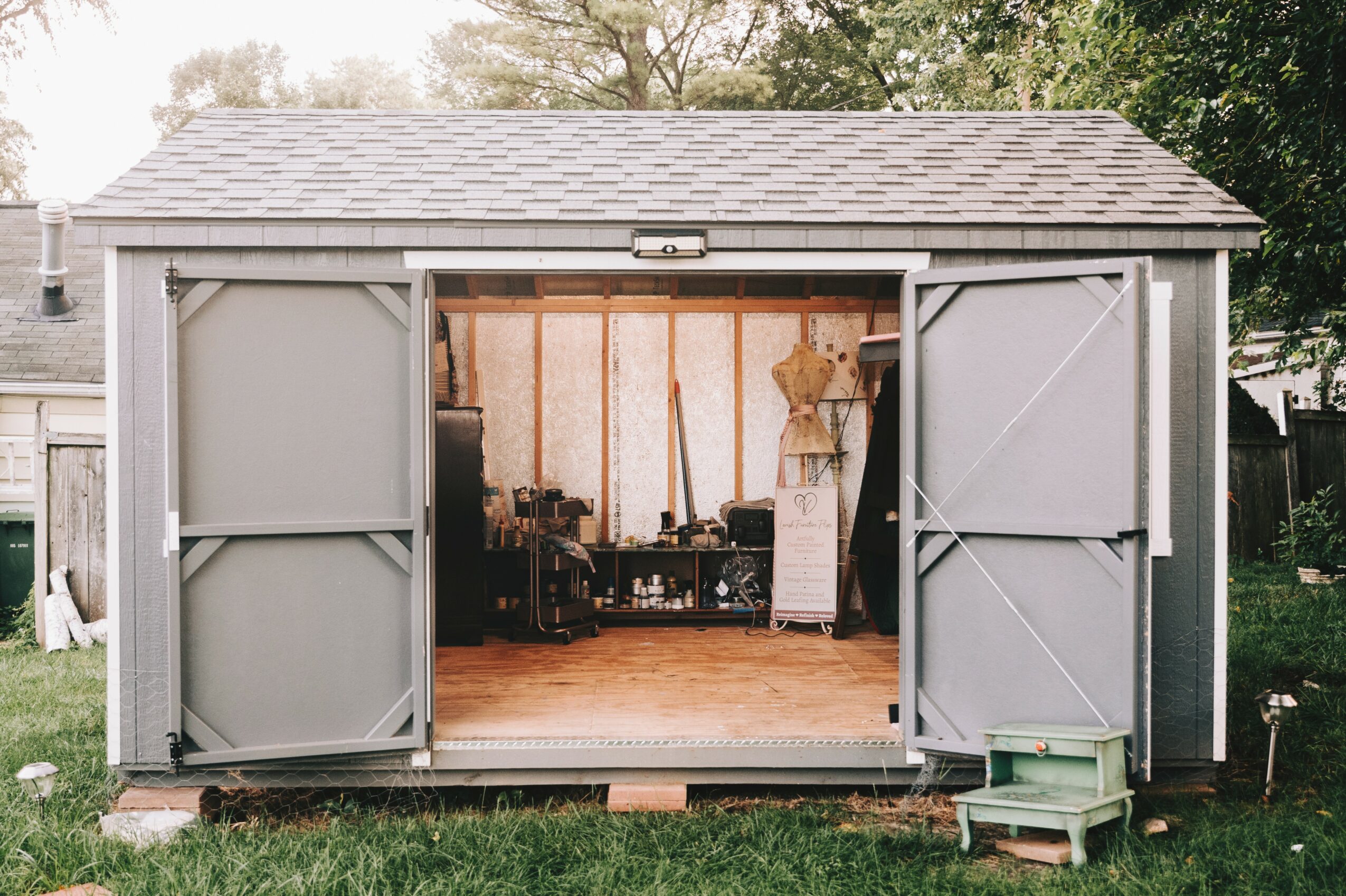 Open garage with tools and equipment inside, a small green step stool outside, and a wooden fence in the background.