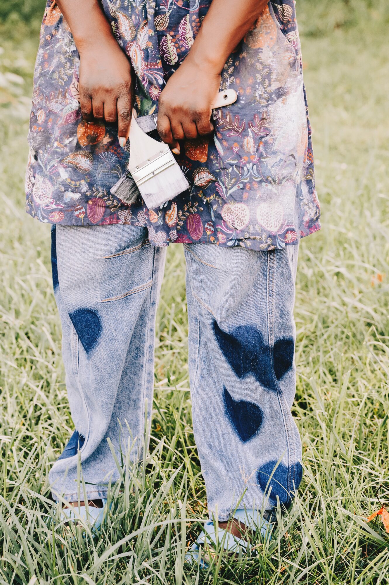 Person standing in grass holding paintbrushes, wearing a floral dress and jeans with heart-shaped patches.