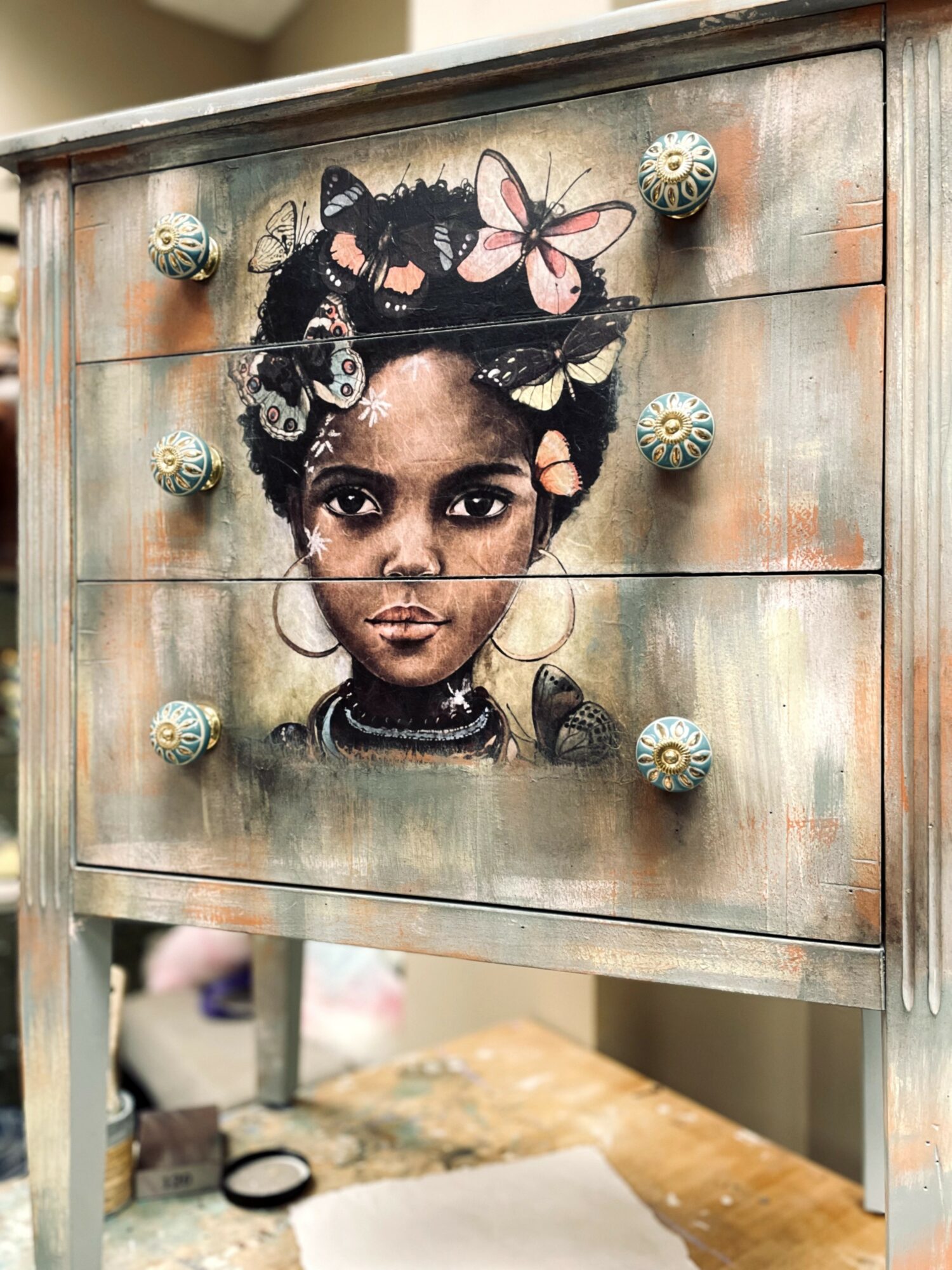 Painting of a young girl with braided hair, adorned with butterflies and flowers, on a wooden dresser.