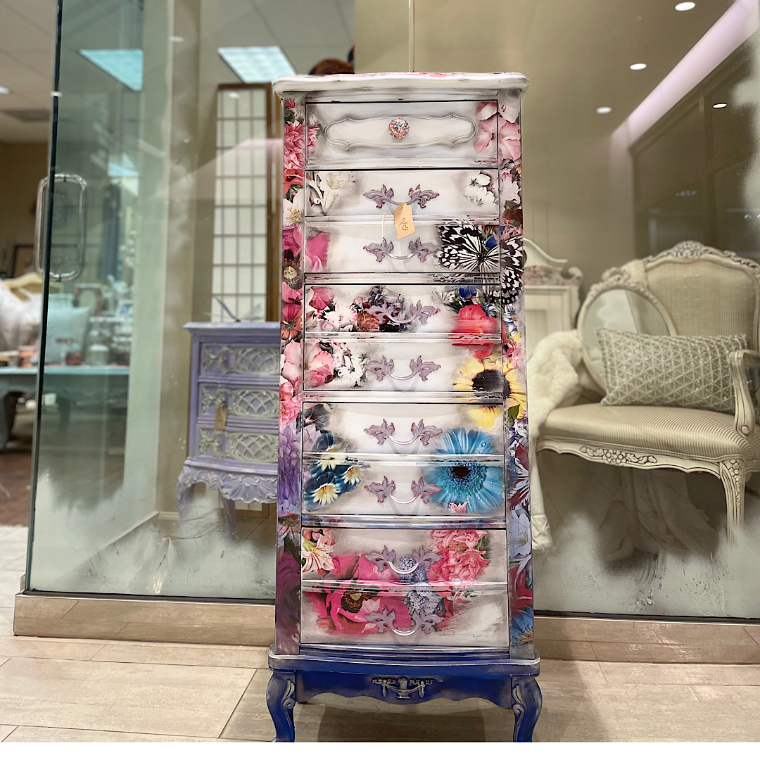 Tall, ornate dresser with floral and butterfly patterns, blue legs, in a room with glass walls and elegant furniture.