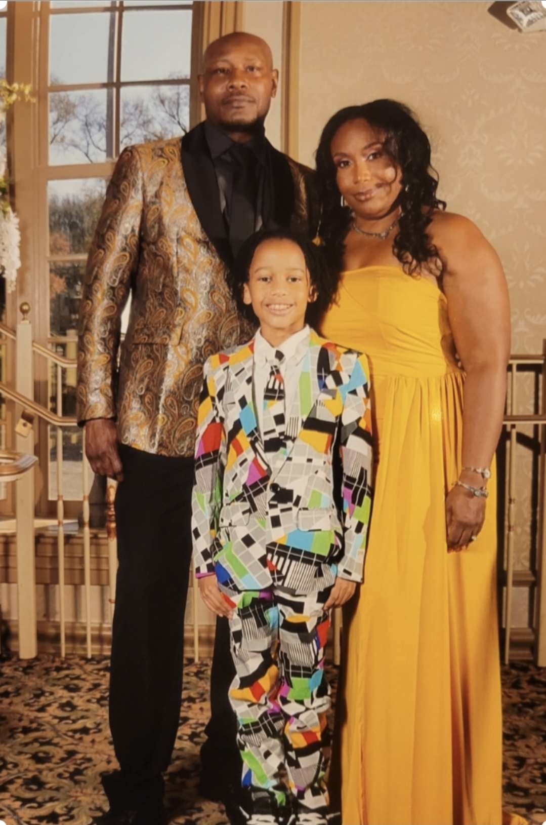 Three people standing indoors, a man in a patterned blazer, a woman in a yellow dress, and a child in a colorful suit.