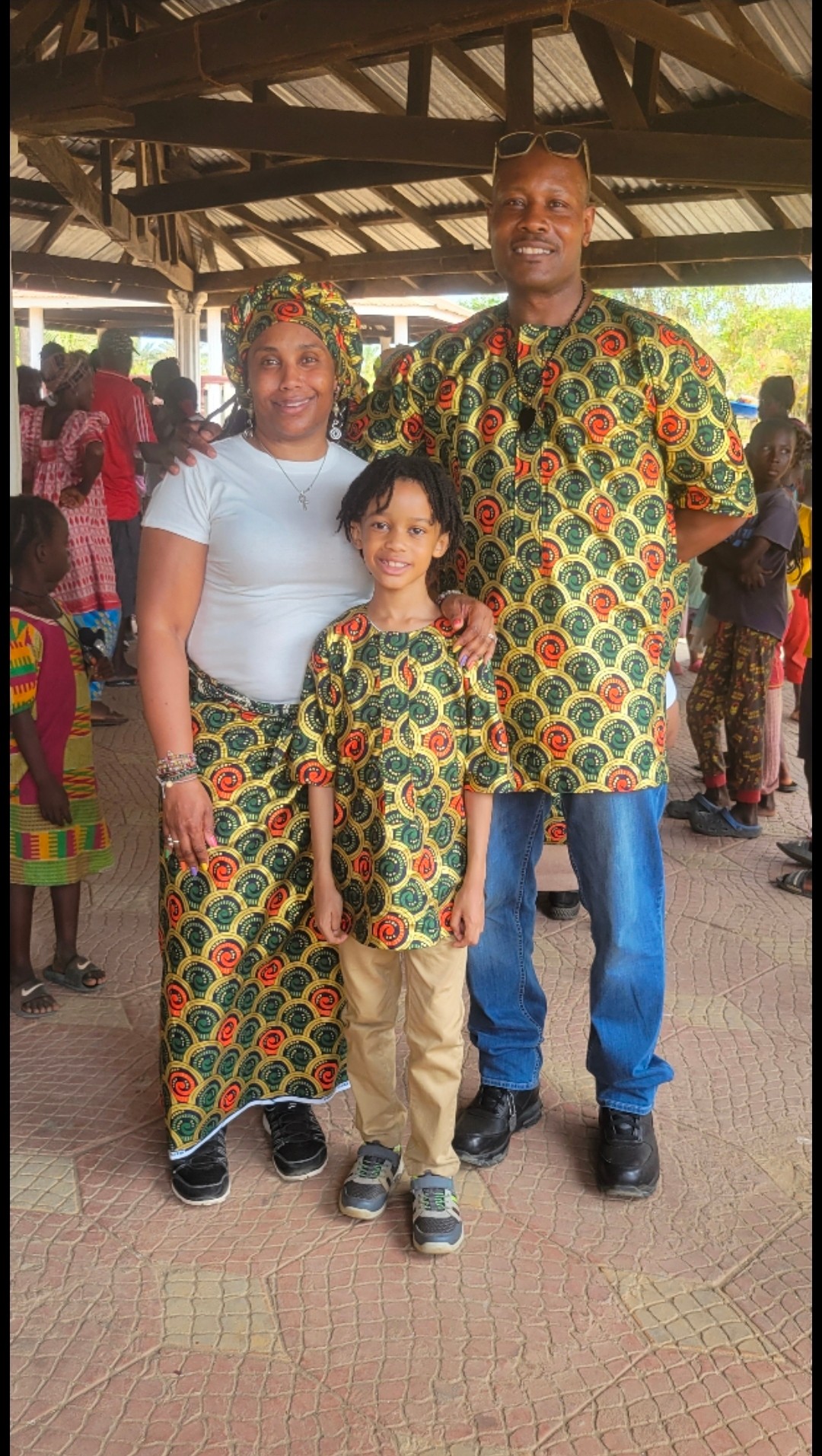 Three people stand outdoors under a wooden roof, wearing matching colorful patterned clothing, with other people in the background.