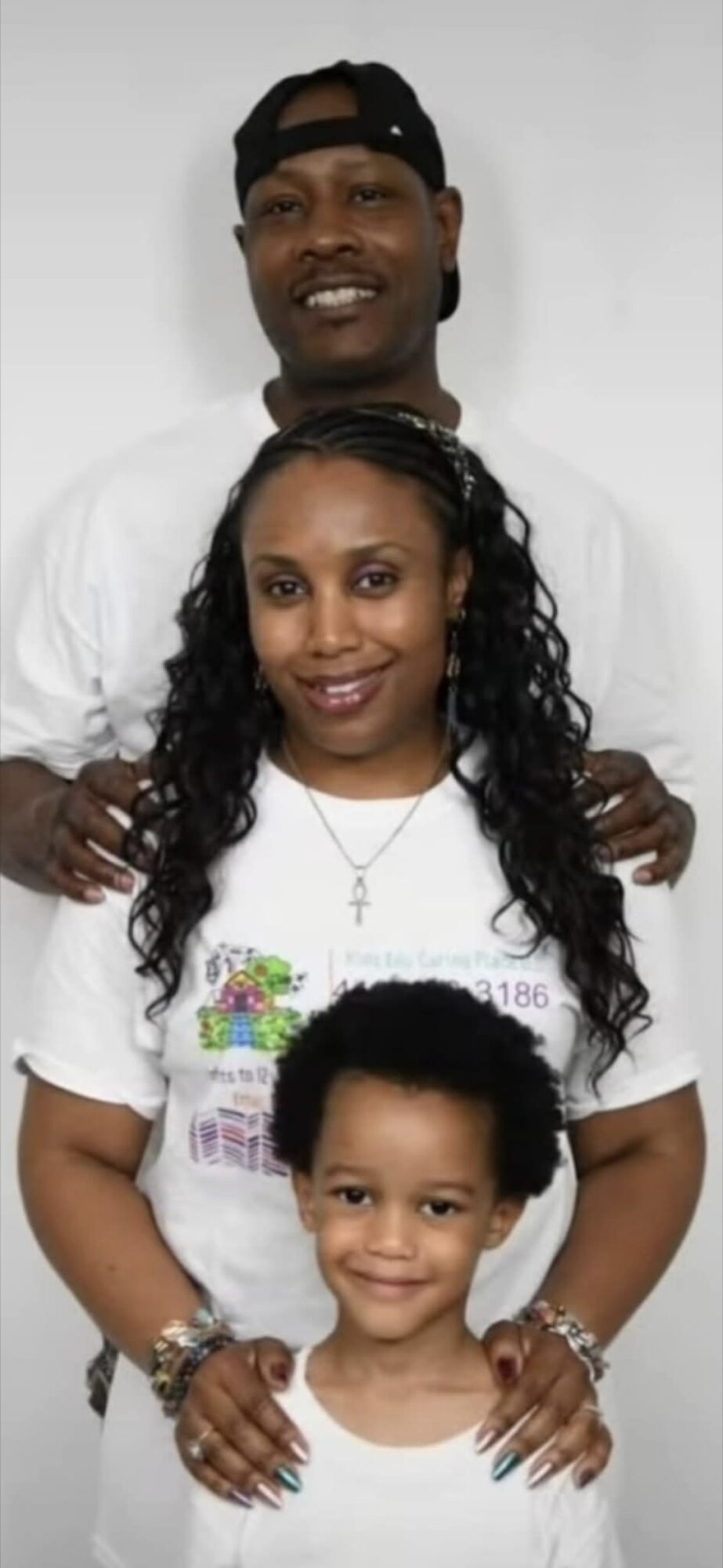 Three people, two adults and one child, smiling and standing close together against a plain background.