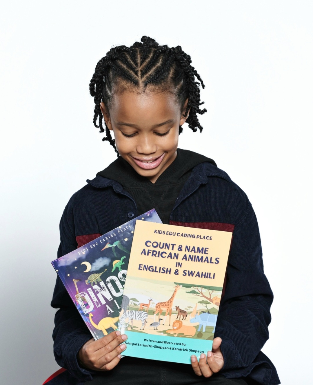 Child holding colorful books, smiling, with braided hair, wearing a dark jacket, against a plain background.