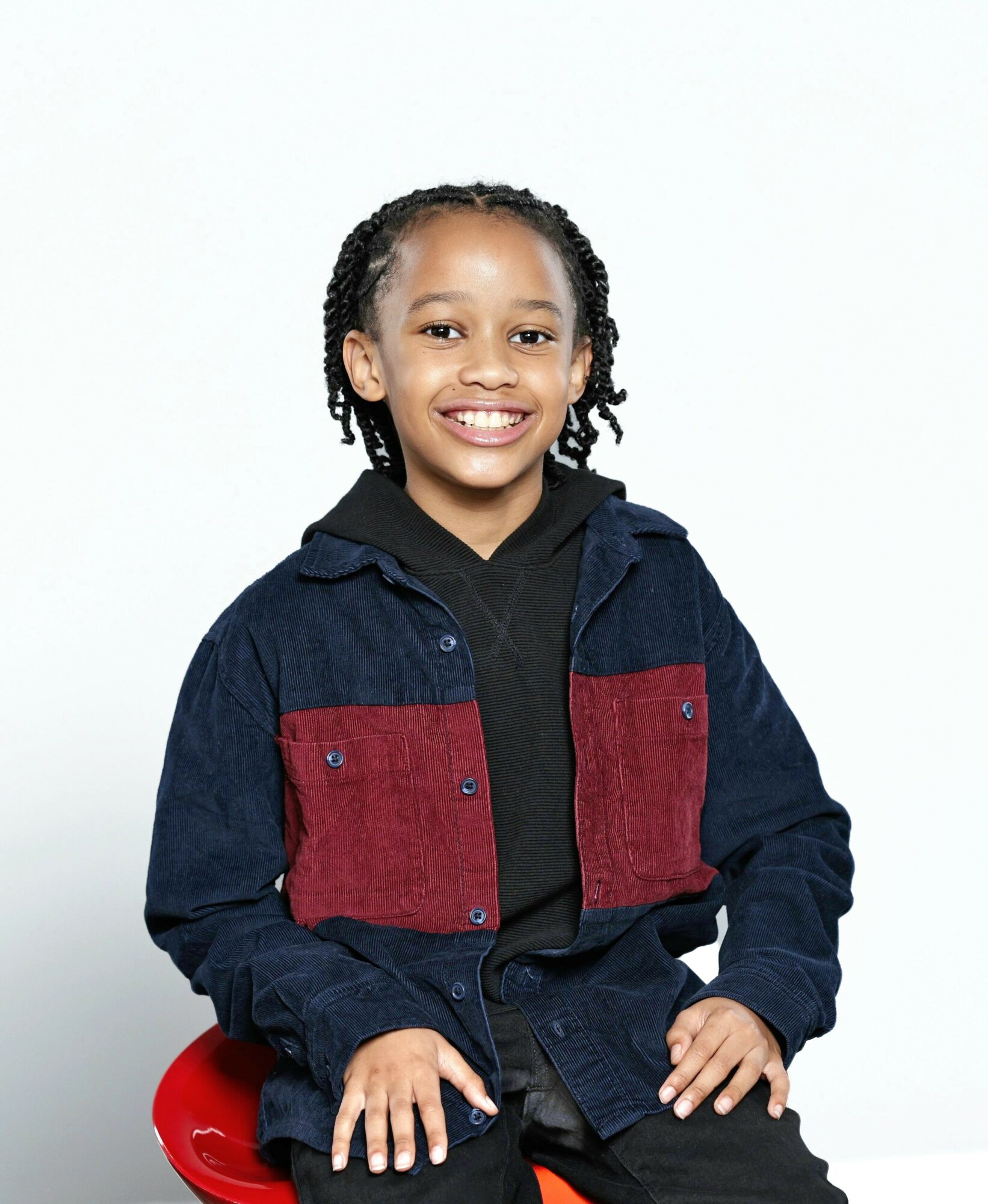 Smiling girl with braided hair wearing a navy and red jacket, black hoodie, sitting on a red chair.