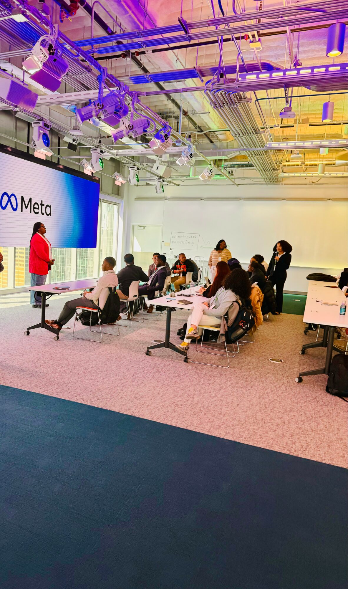 People seated in a conference room with a presenter standing near a large screen displaying 'Meta'.