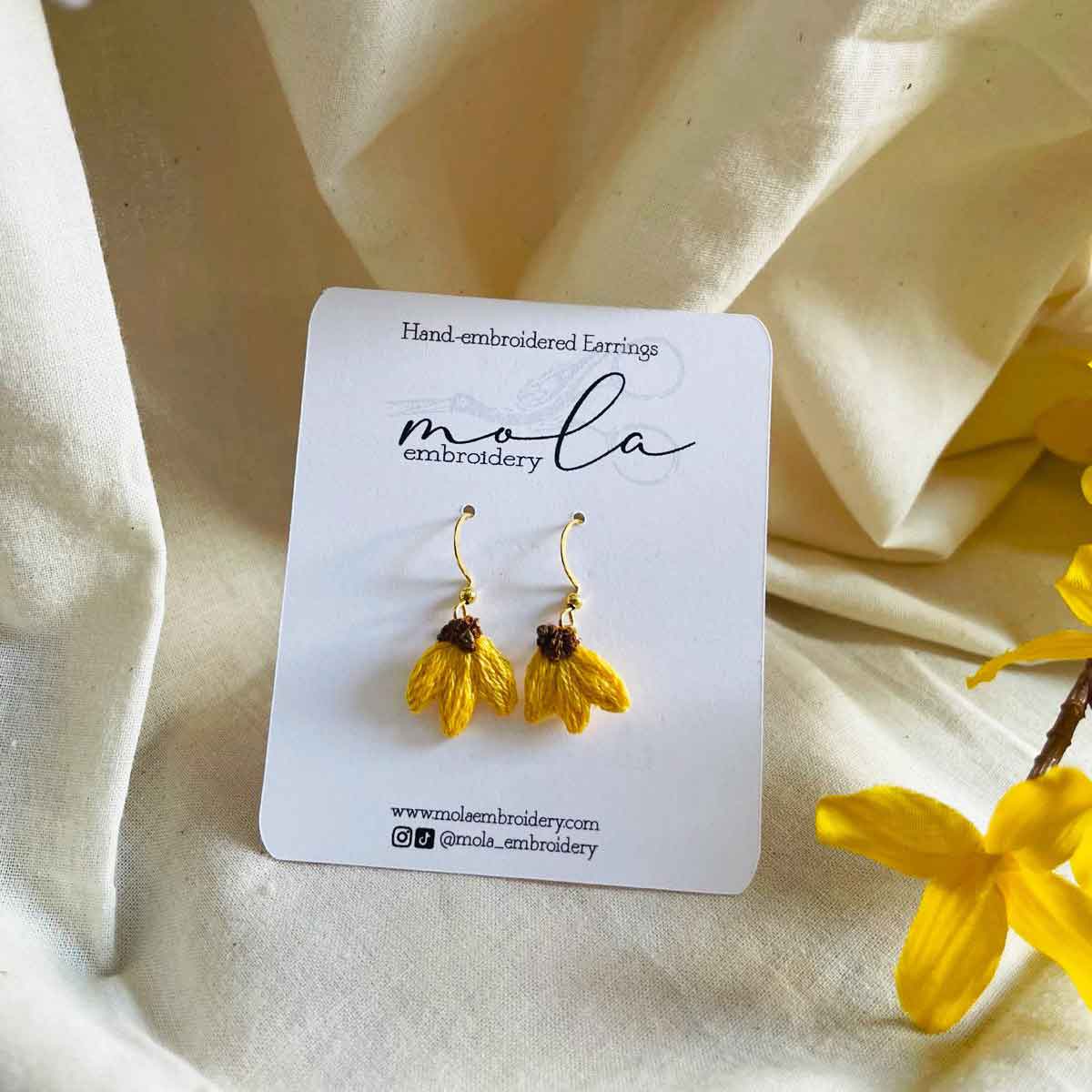 Yellow tassel earrings with gold hooks on a white card, surrounded by cream-colored fabric and yellow flowers.