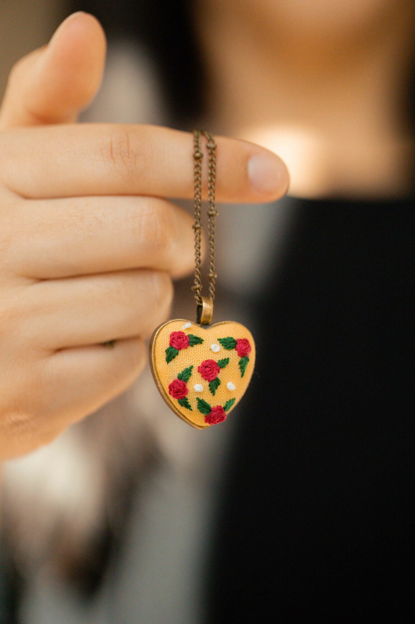 Person holds a heart-shaped pendant necklace with a floral pattern, fingers gently grasping the chain.