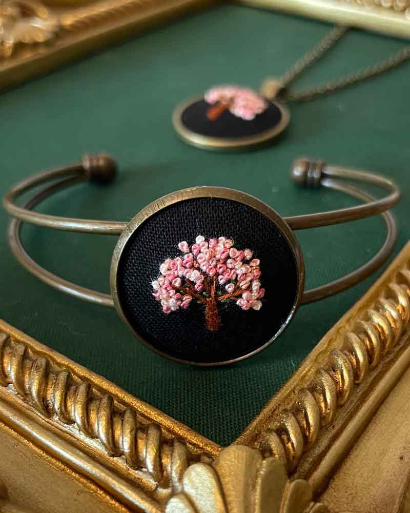 Bracelet with circular pendant featuring a pink flowering tree on black background, displayed on green fabric with gold frame.