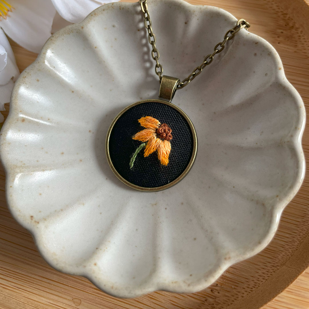 Pendant with embroidered orange and brown flower on black background, on a gold chain, in a white ceramic dish.