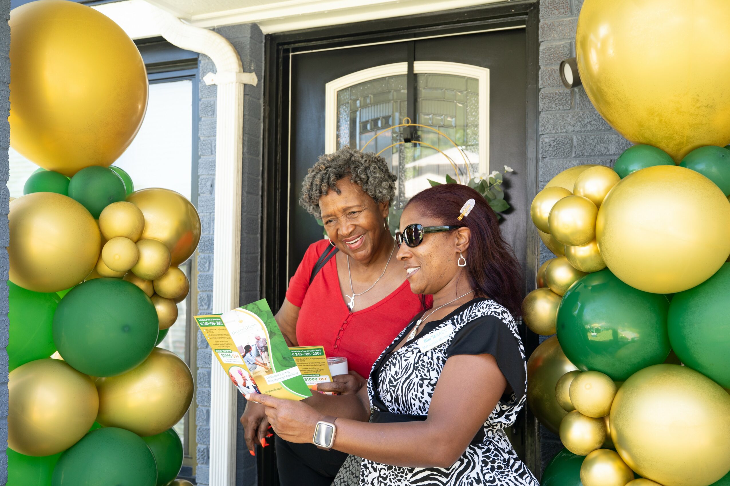 Two women stand outside near balloon decorations, looking at a colorful brochure or flyer together, smiling.