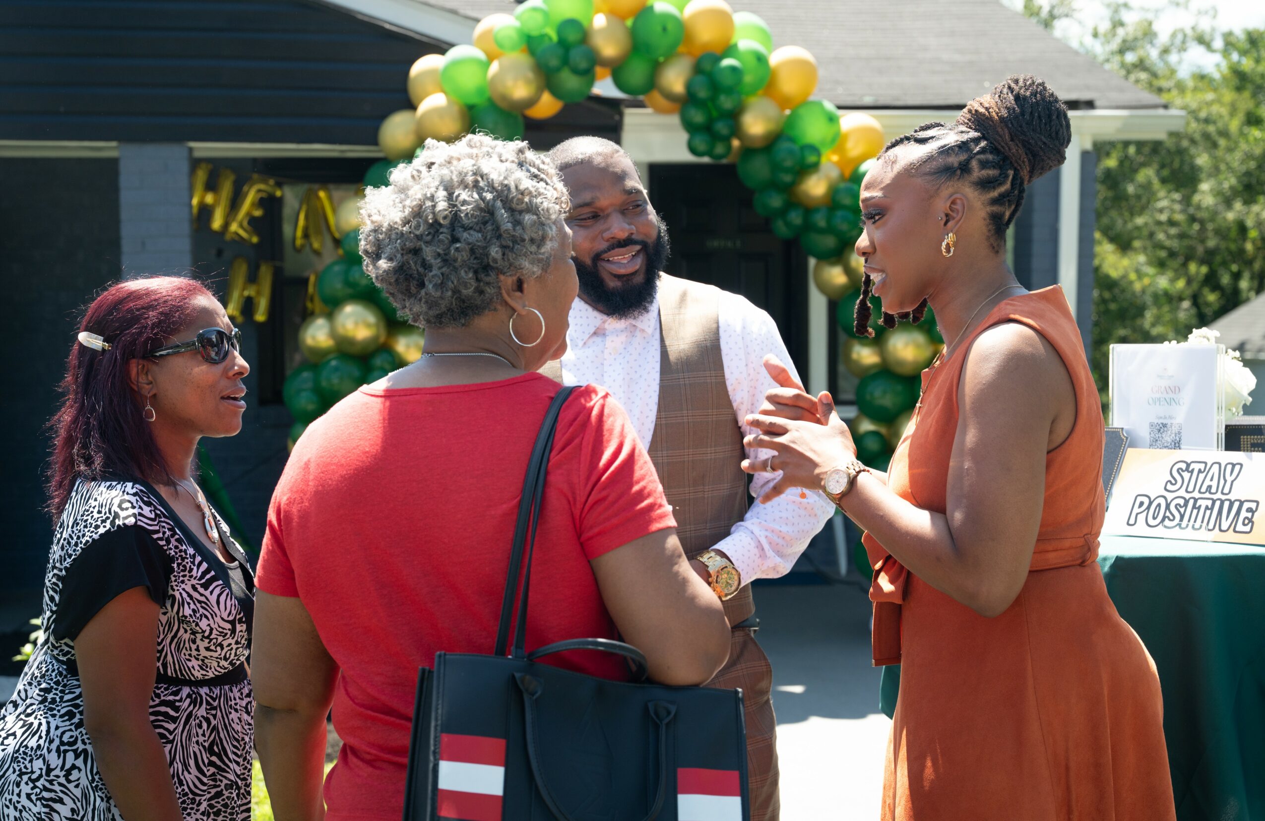 Four people engaged in conversation outdoors near a balloon arch, with a sign reading 'Stay Positive' in the background.