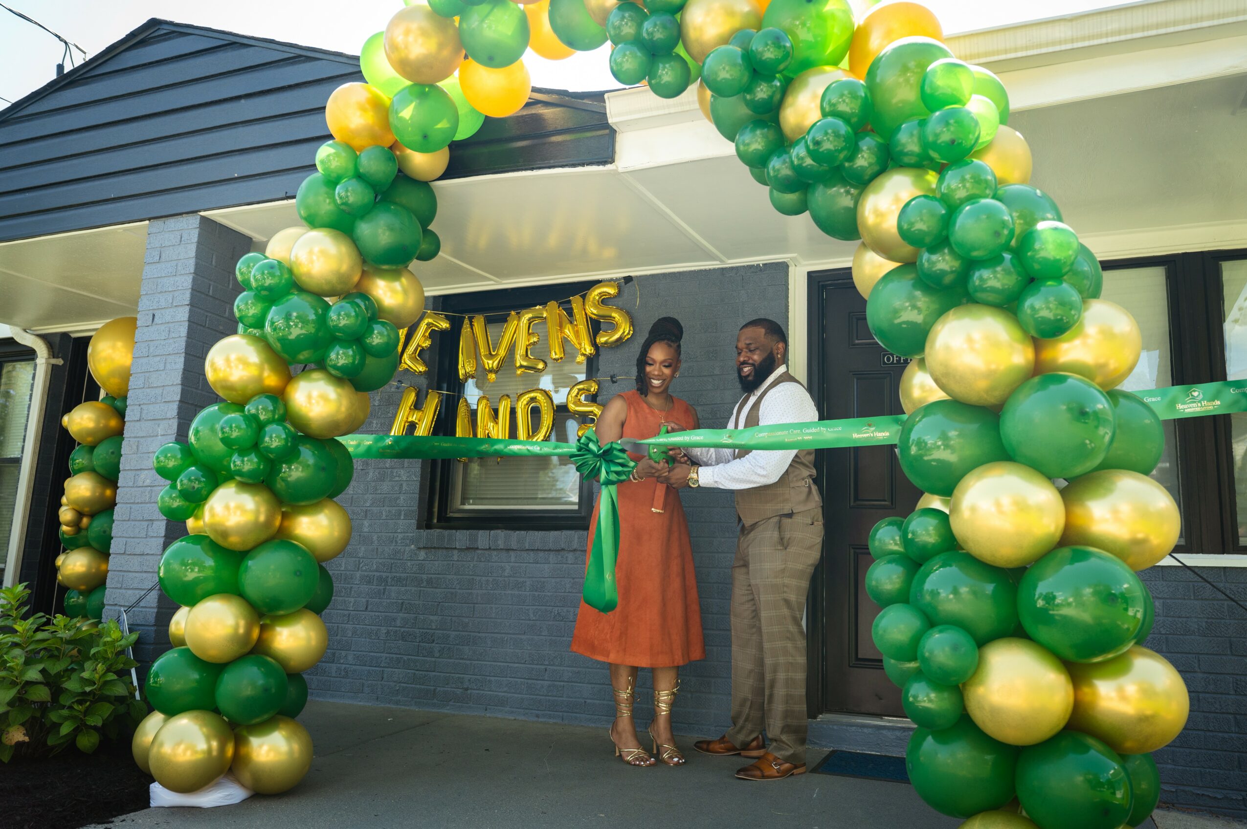Three people stand under a green and gold balloon arch outside a building, holding a ribbon for a ribbon-cutting event.