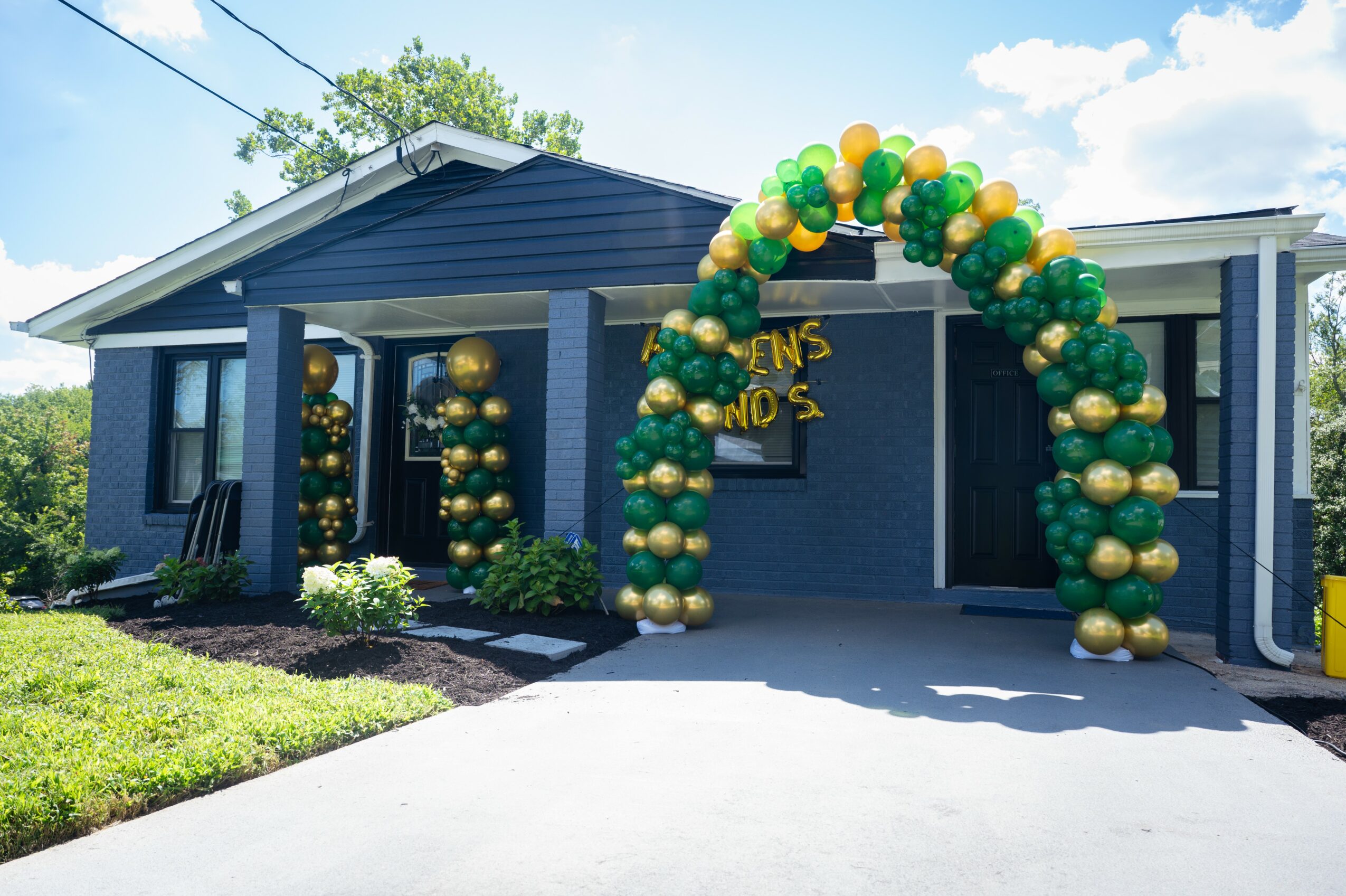 House decorated with green and gold balloons forming an arch and columns, with a sign partially visible on the wall.