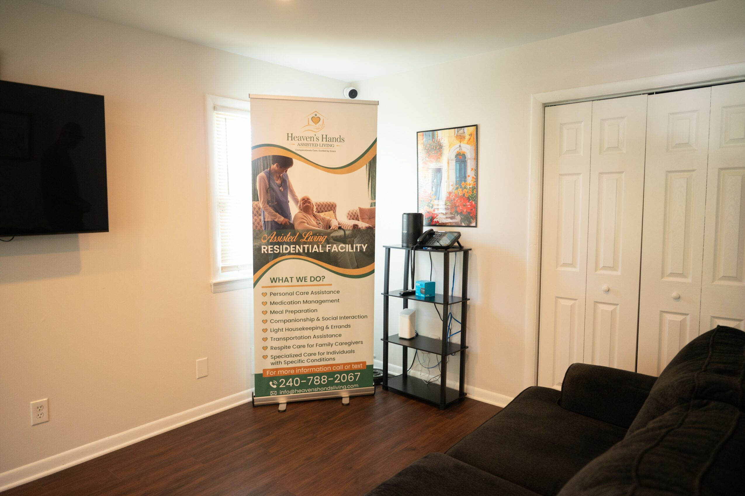 Living room with window, banner, black shelf, artwork, and black couch, wood floor, white walls, and closet doors.