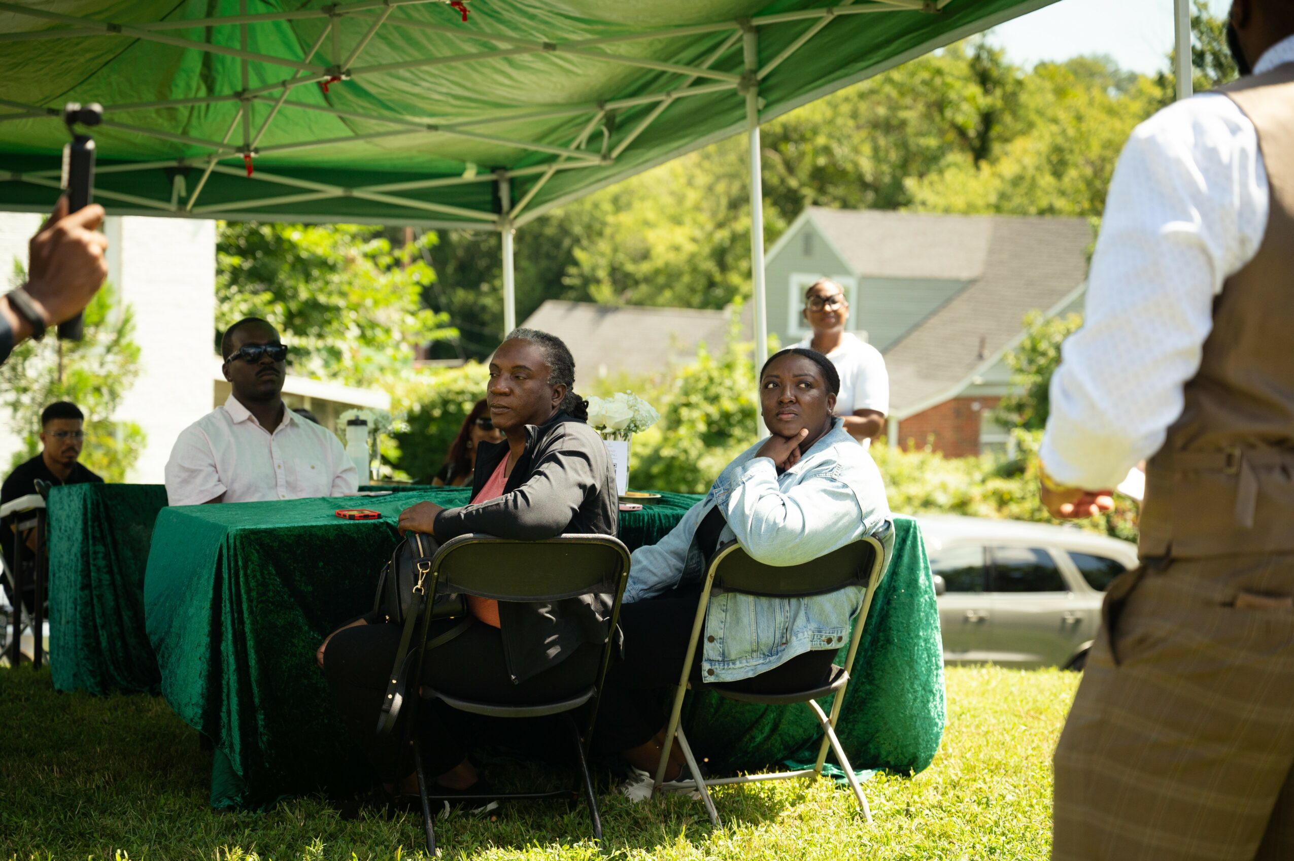 Group of people seated at a table outdoors under a green canopy, with trees and houses in background.
