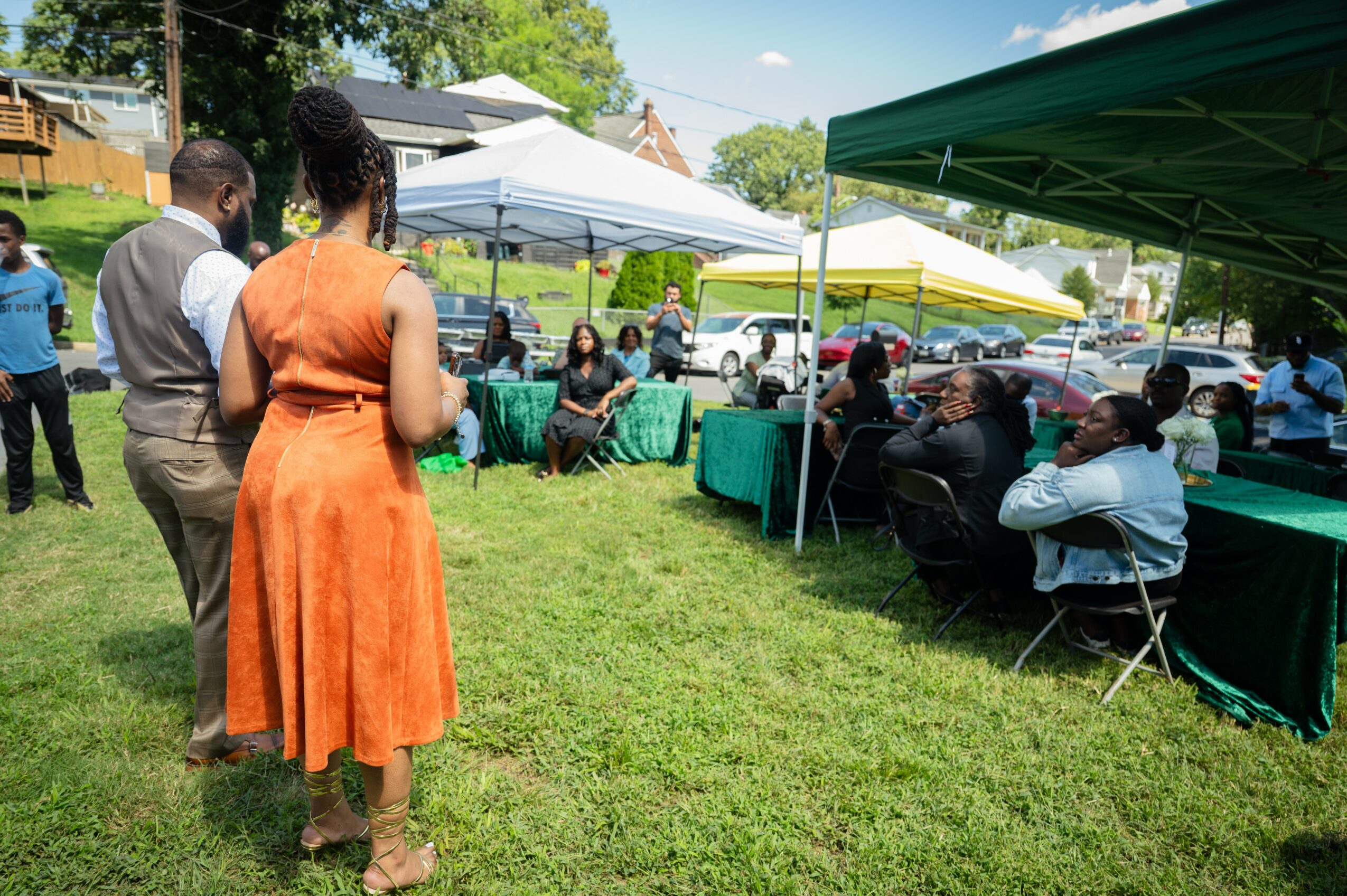 People gathered outdoors under tents, sitting at tables, and standing on grass during daytime event.