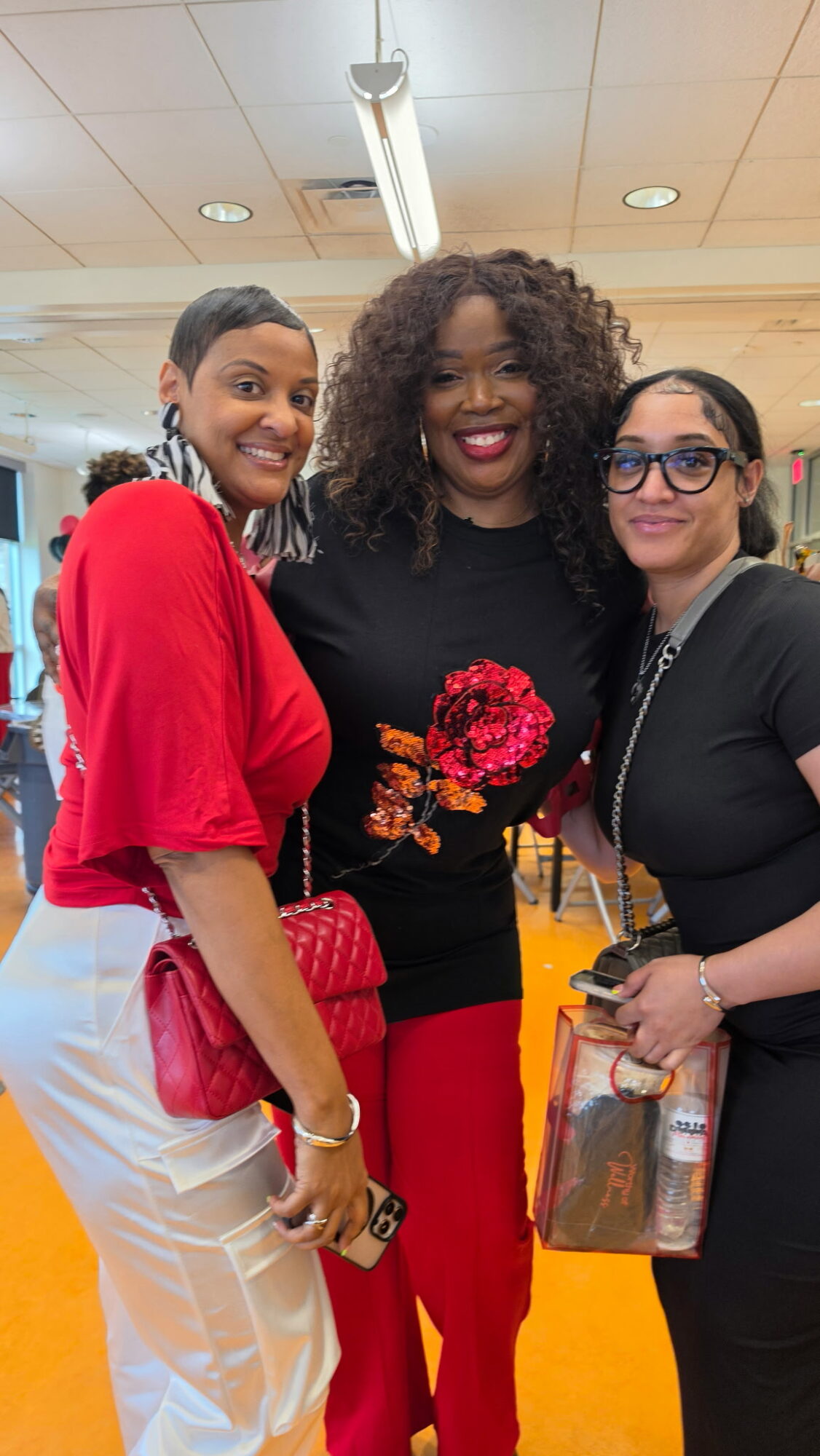 Three women smiling indoors, one holding a purse, in casual attire, with a ceiling light above.