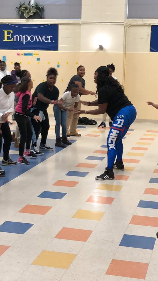 Group of children and an adult in a gymnasium participating in an activity, with colorful tiles on the floor and a banner in the background.