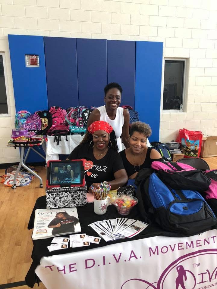 Three women sitting at a table with bags, a laptop, and flyers, in front of a blue wall, smiling.